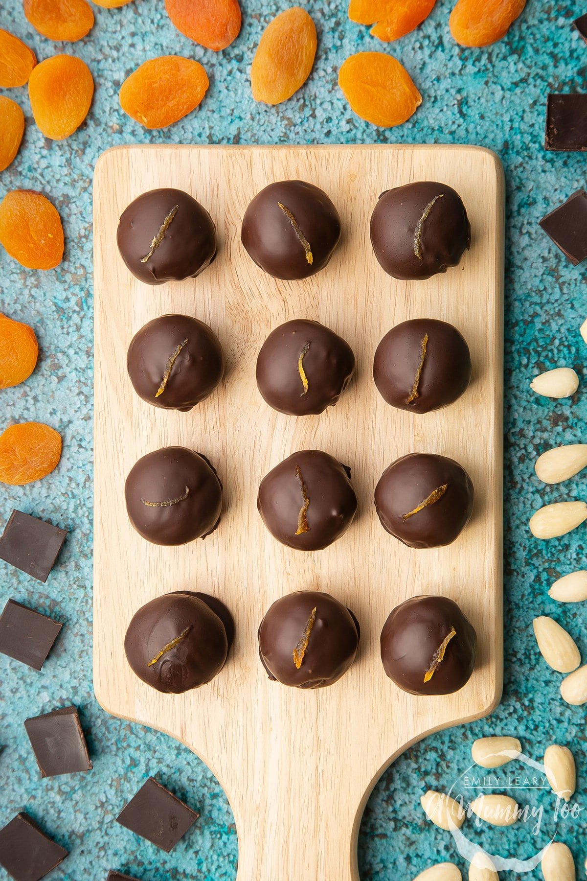 12 chocolate apricot balls arranged on a wooden board. Dried apricots, blanched almonds and squares of dark chocolate surround the board.