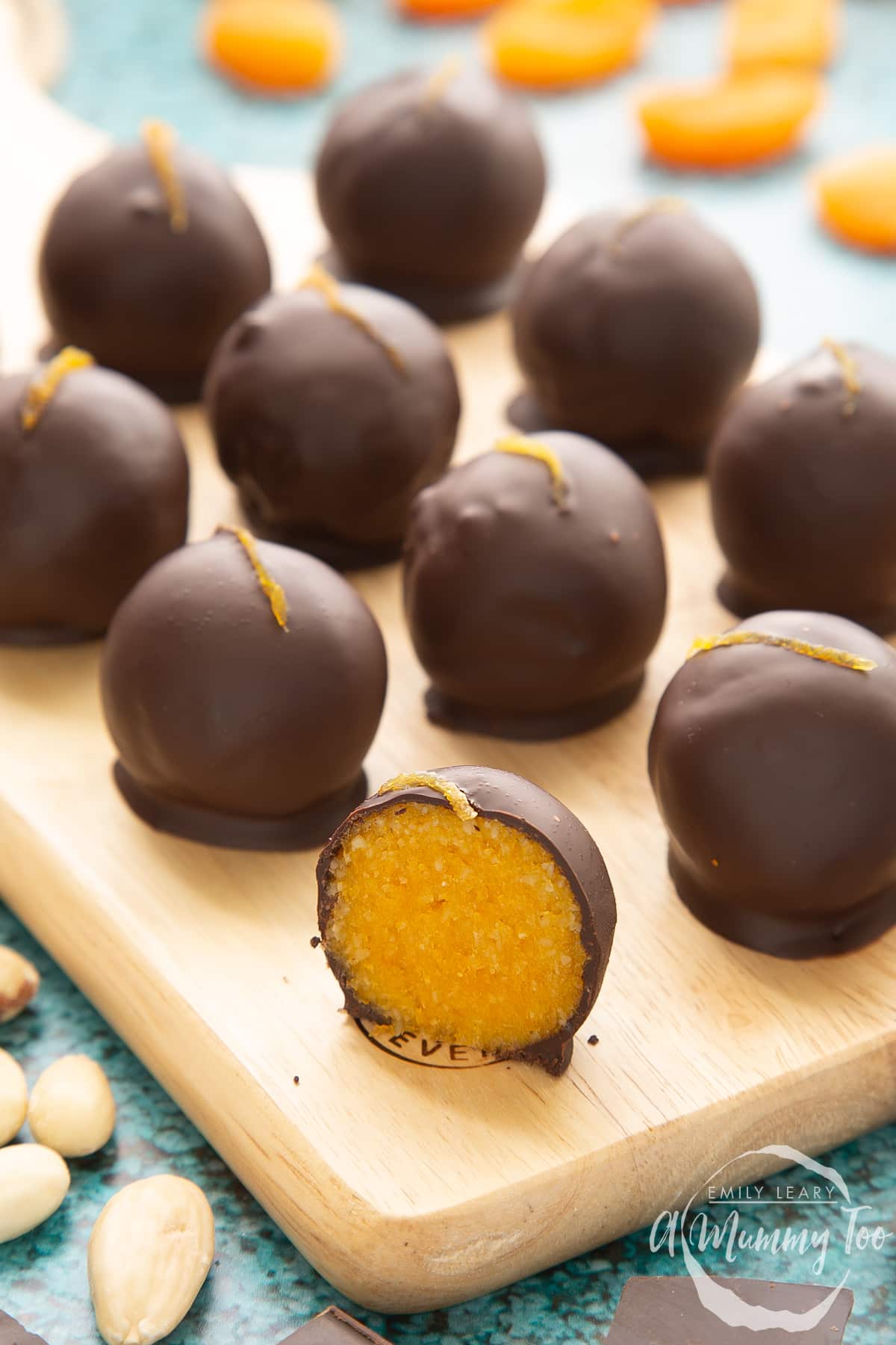 Chocolate apricot balls arranged on a wooden board. Dried apricots, blanched almonds and squares of dark chocolate surround the board. In the foreground sits half a ball, revealing the bright orange filling.