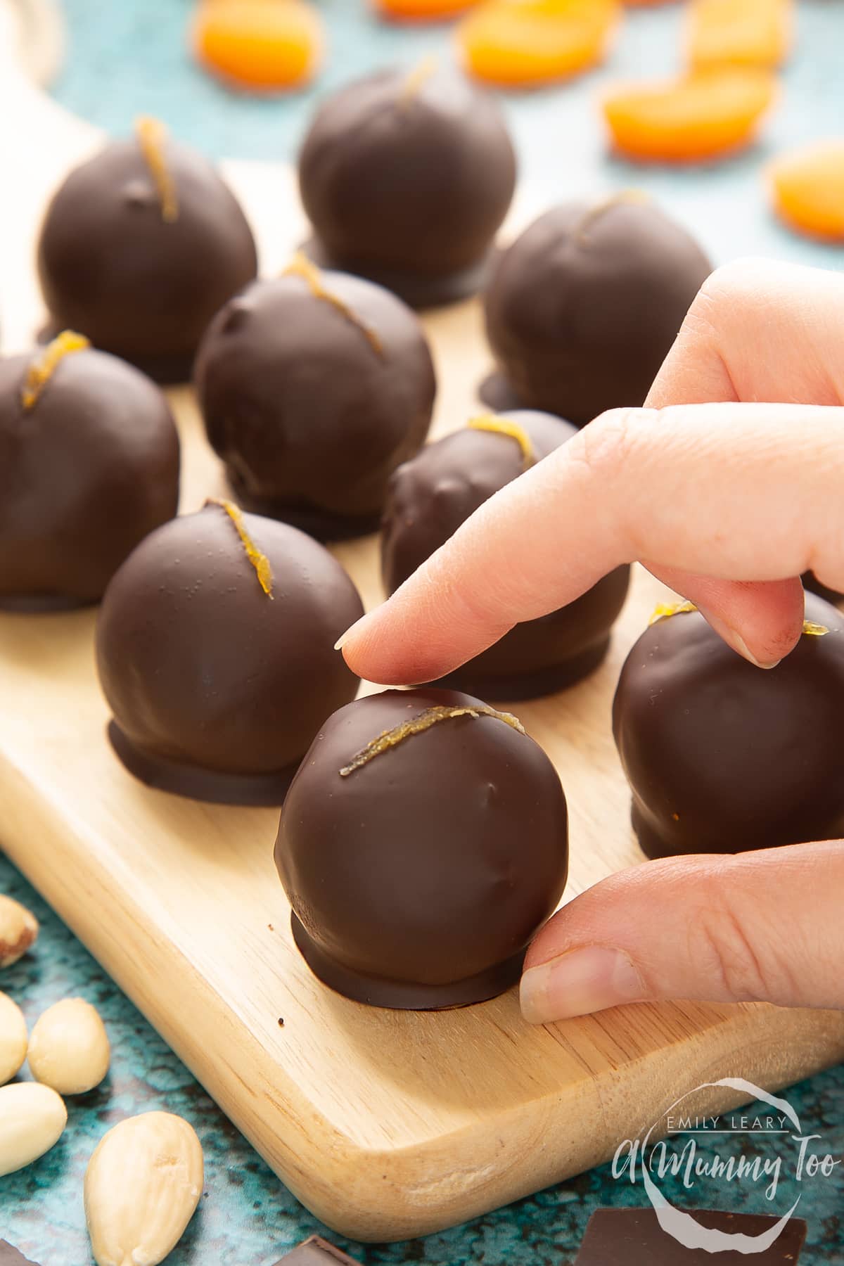 Chocolate apricot balls arranged on a wooden board. Dried apricots, blanched almonds and squares of dark chocolate surround the board. A hand reaches in to take one. 