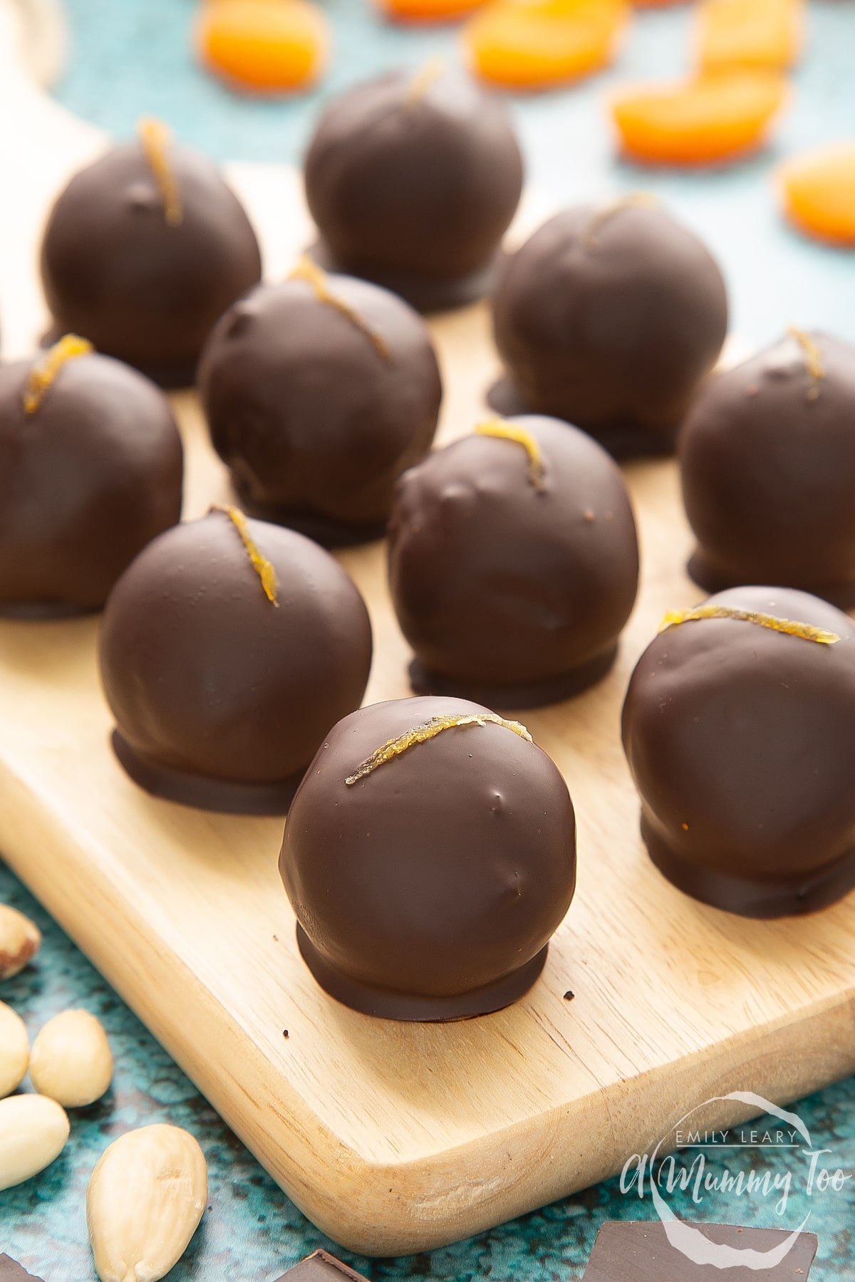 Chocolate apricot balls arranged on a wooden board. Dried apricots, blanched almonds and squares of dark chocolate surround the board. 