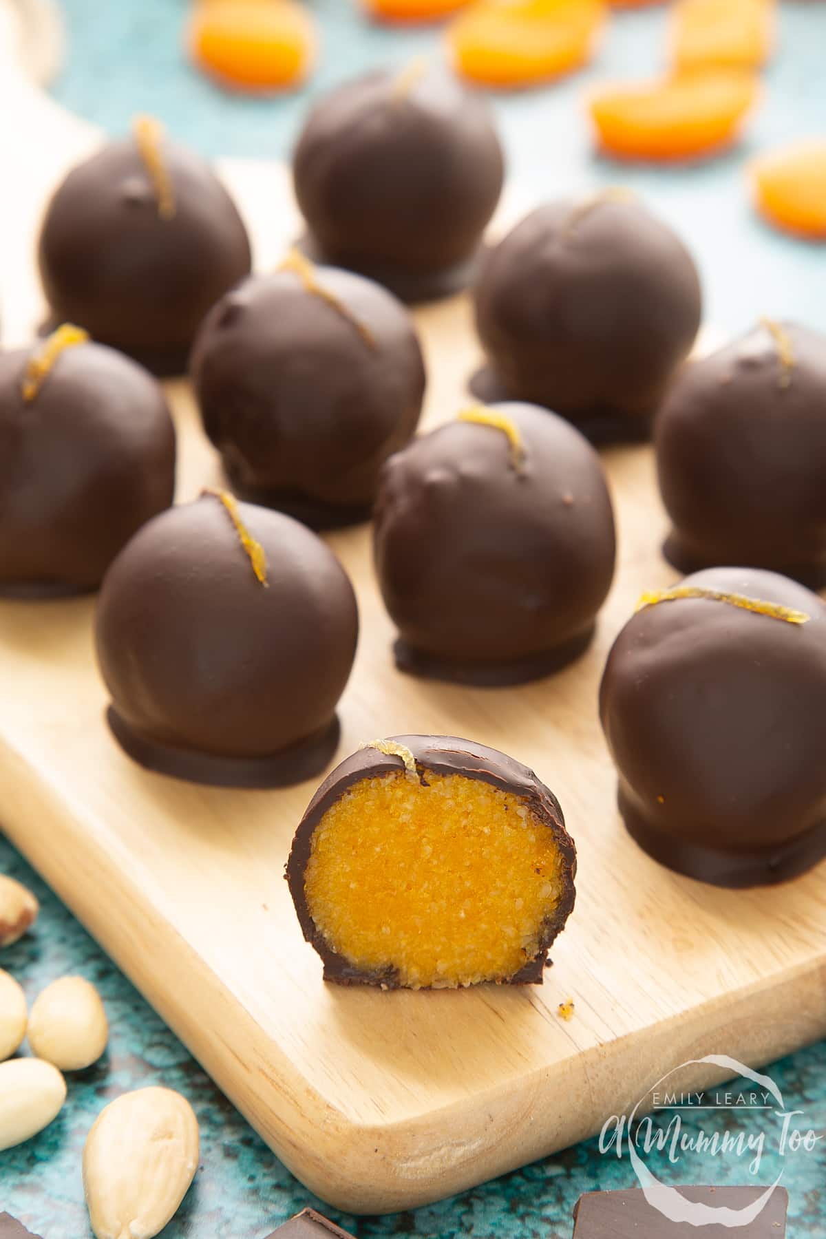 Chocolate apricot balls arranged on a wooden board. In the foreground sits half a ball, revealing the bright orange filling.