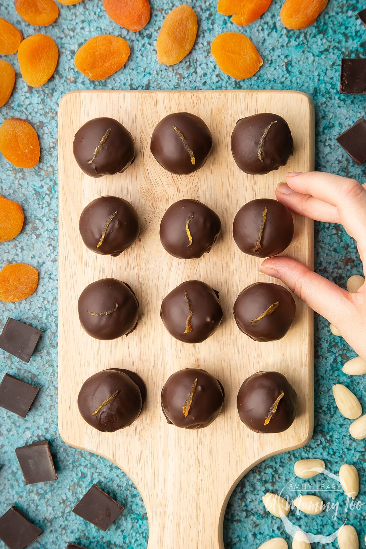 12 chocolate apricot balls arranged on a wooden board. A hand reaches in to take one. Dried apricots, blanched almonds and squares of dark chocolate surround the board.