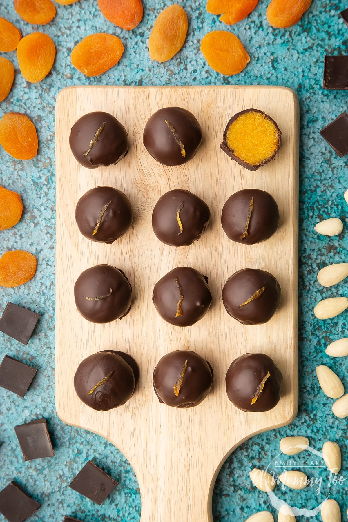 12 chocolate apricot balls arranged on a wooden board. One has been cut in half, revealing the bright orange apricot filling. Dried apricots, blanched almonds and squares of dark chocolate surround the board.