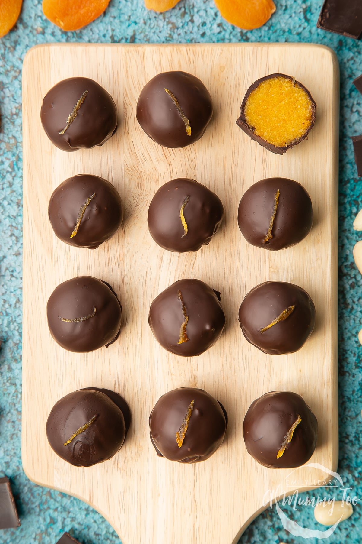 12 chocolate apricot balls arranged on a wooden board, with ingredients around the board. One has been cut in half, revealing the bright orange apricot filling. 