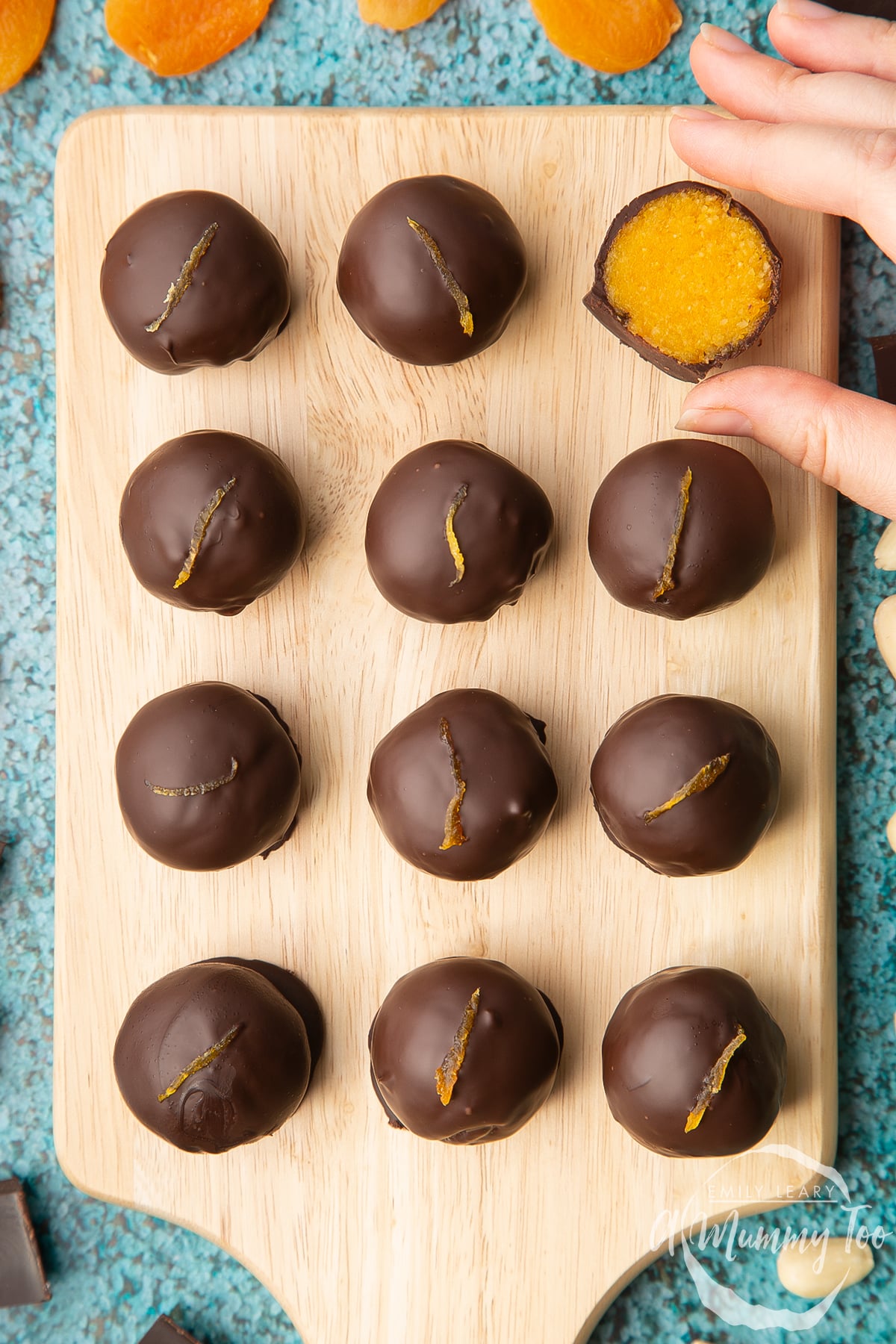 12 chocolate apricot balls arranged on a wooden board. A hand reaches to take one that has been cut in half, revealing the bright orange apricot filling. 