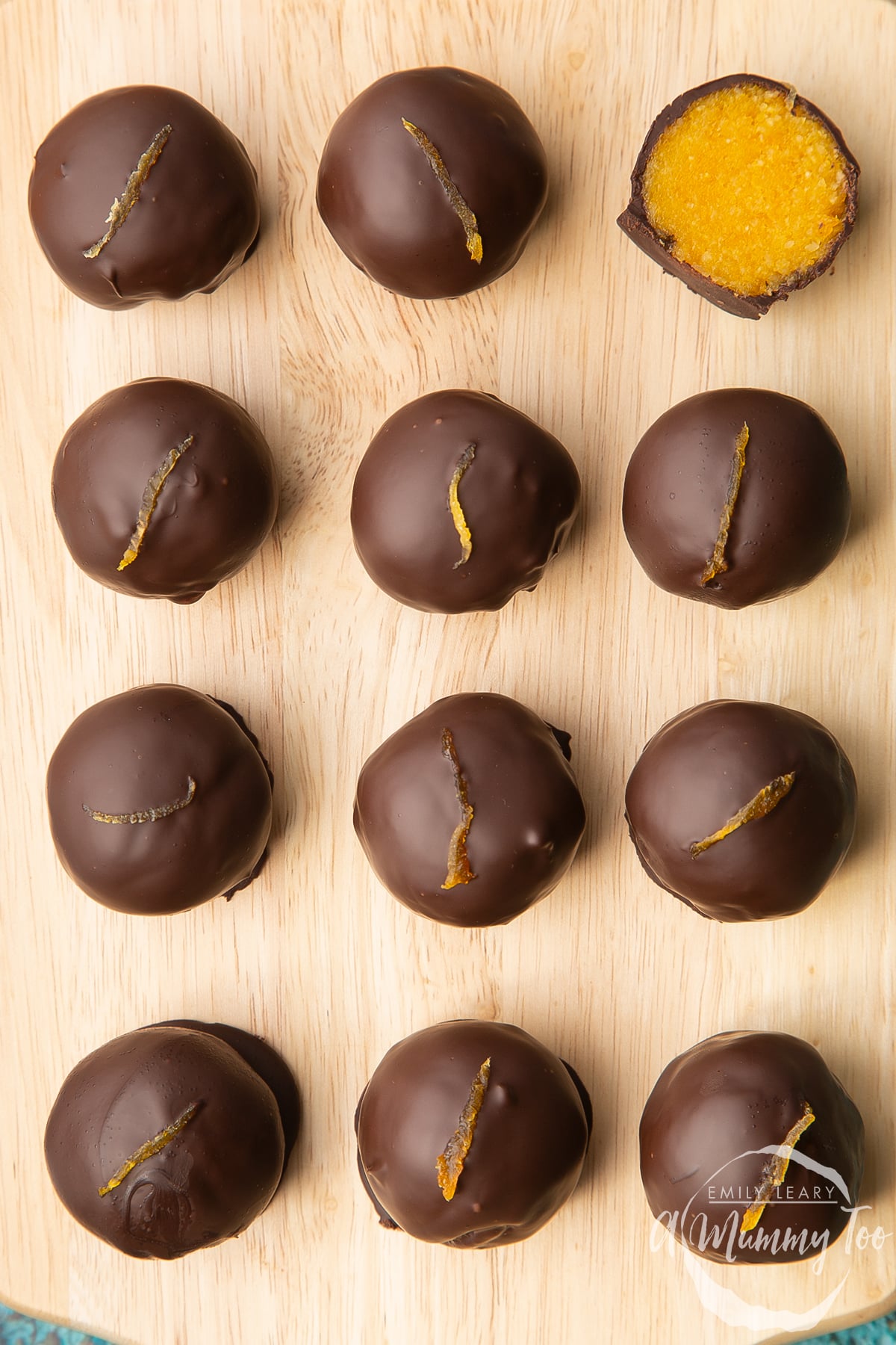 12 chocolate apricot balls arranged on a wooden board. One has been cut in half, revealing the bright orange apricot filling. 