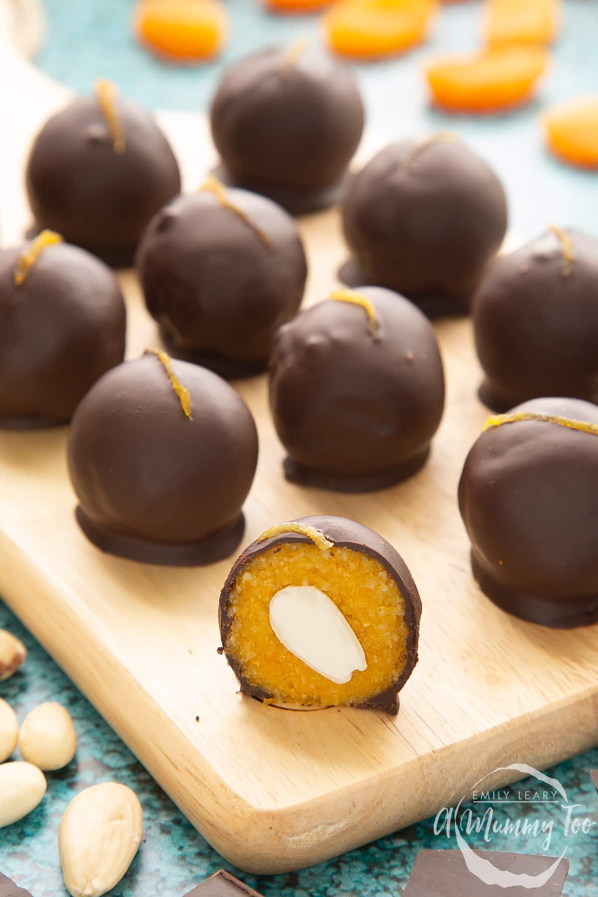 Chocolate apricot balls arranged on a wooden board. In the foreground sits half a ball, revealing the bright orange filling. An almond half sits in the centre of the filling.