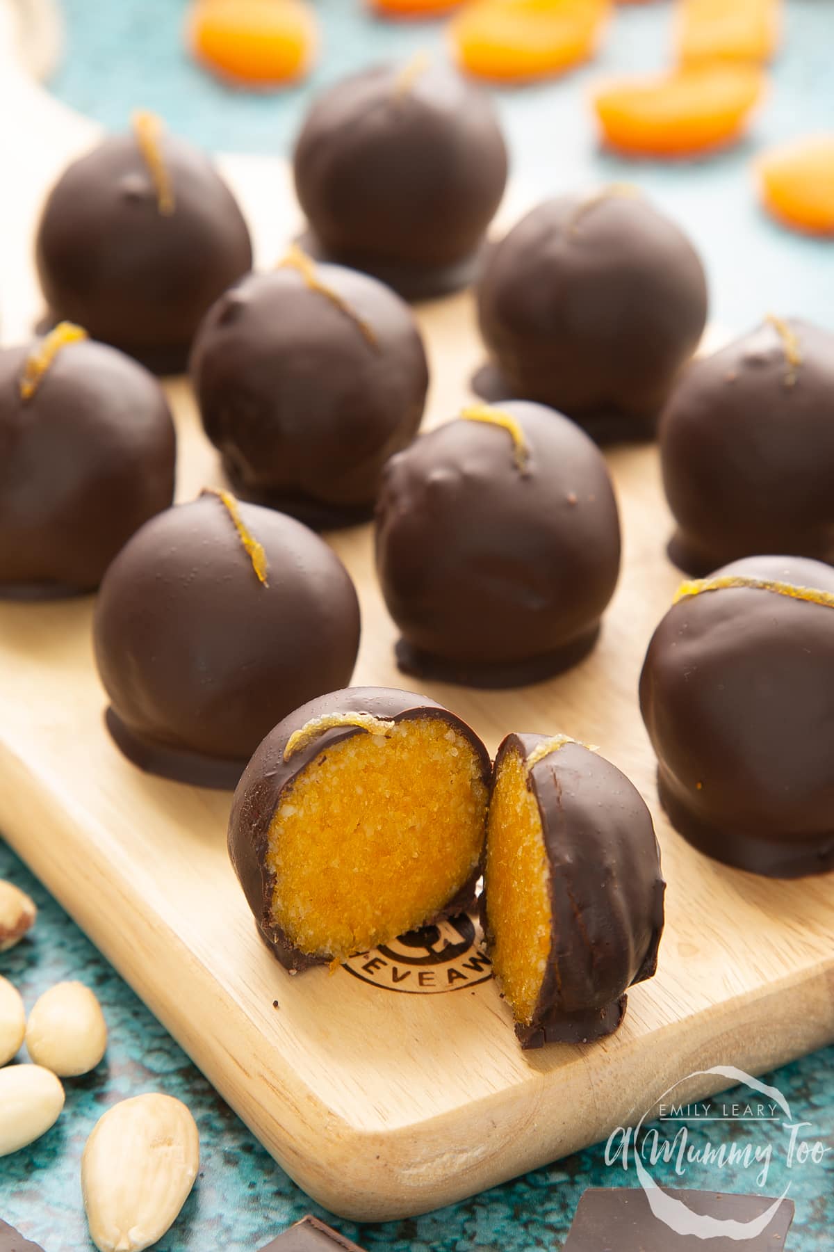 Chocolate apricot balls arranged on a wooden board. Dried apricots, blanched almonds and squares of dark chocolate surround the board. The ball in the foreground has been cut in two, revealing the bright orange filling.