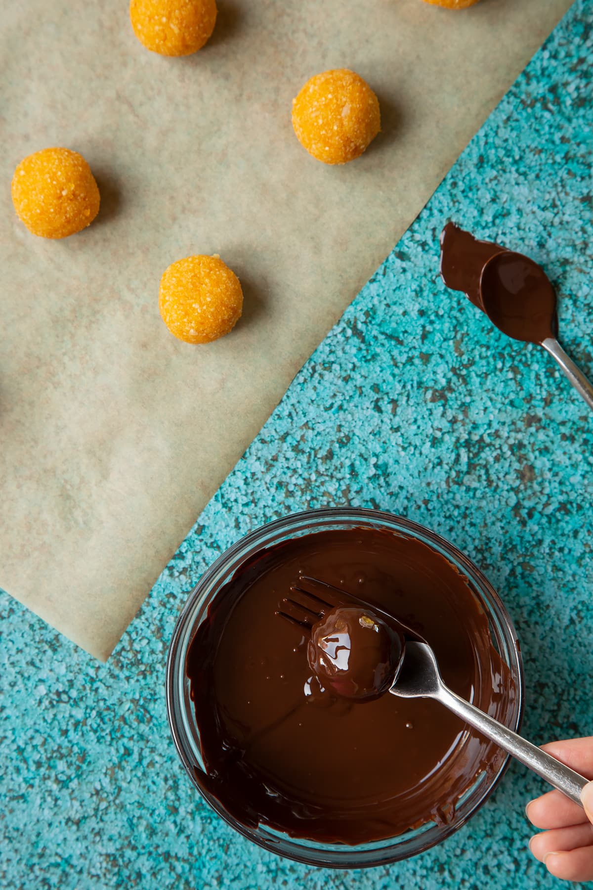 Balls made from blended, apricots and nuts. A small mixing bowl containing melted dark chocolate. A fork lifts an apricot ball coated in chocolate from the bowl.