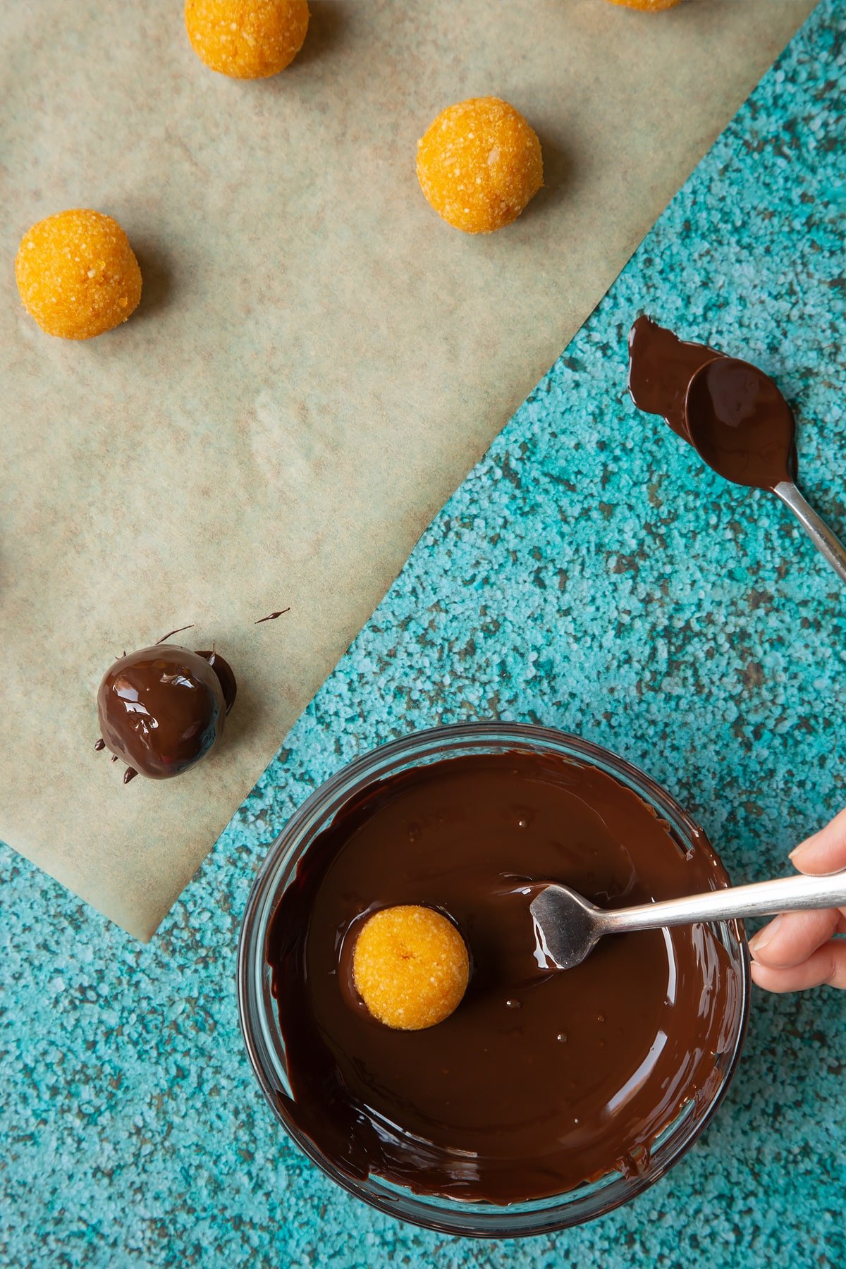 Balls made from blended, apricots and nuts. A small mixing bowl containing melted dark chocolate. A fork lowers an apricot ball into the chocolate.