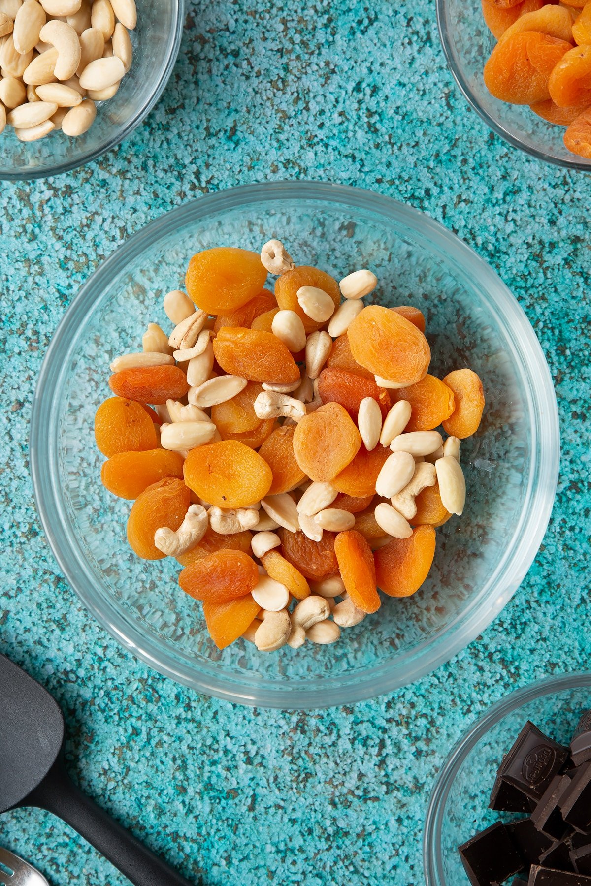 A mixing bowl containing blanched almonds, apricots and a few cashews. Ingredients to make chocolate apricot balls surround the ball.