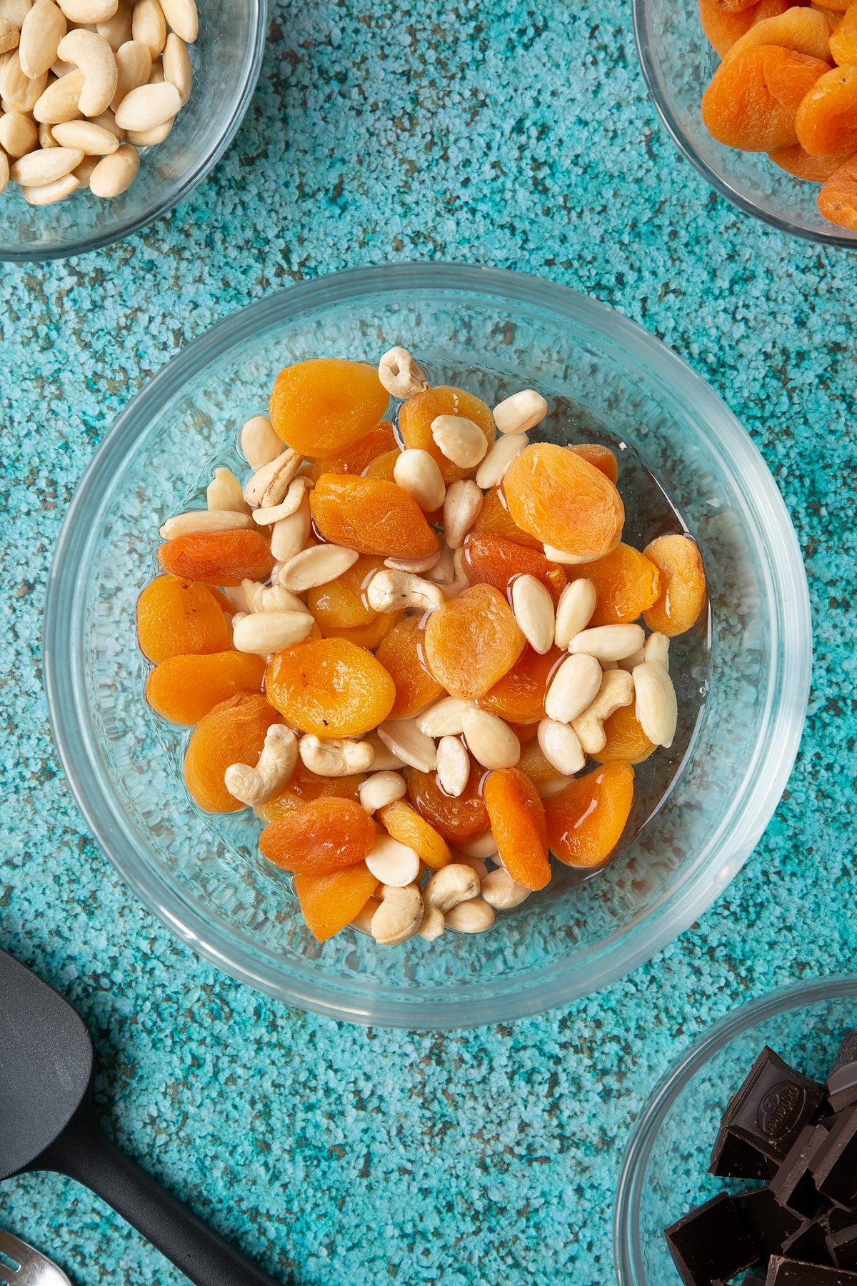 A mixing bowl containing blanched almonds, apricots and a few cashews soaking in hot water. Ingredients to make chocolate apricot balls surround the ball.