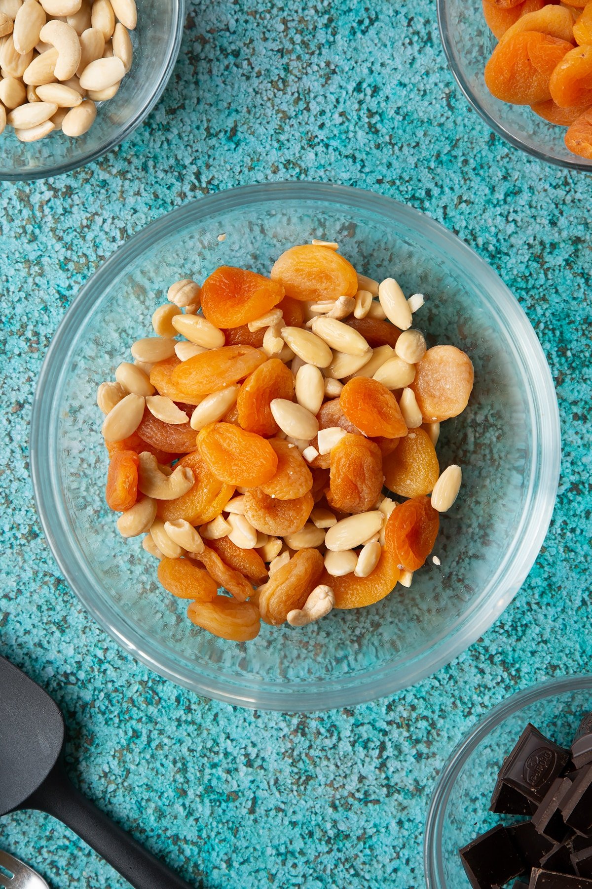 A mixing bowl containing soaked blanched almonds, apricots and a few cashews. Ingredients to make chocolate apricot balls surround the ball.