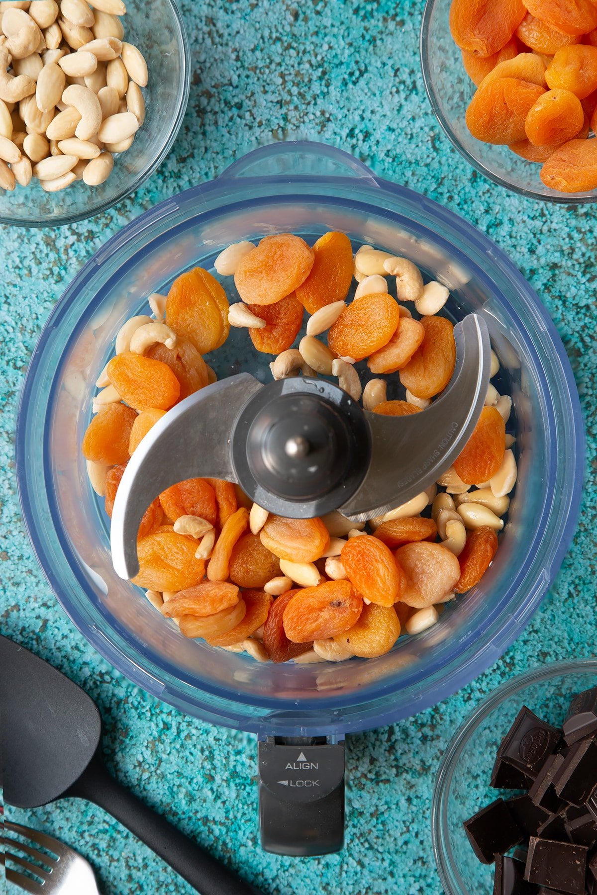 A blender containing soaked blanched almonds, apricots and a few cashews. Ingredients to make chocolate apricot balls surround the blender.