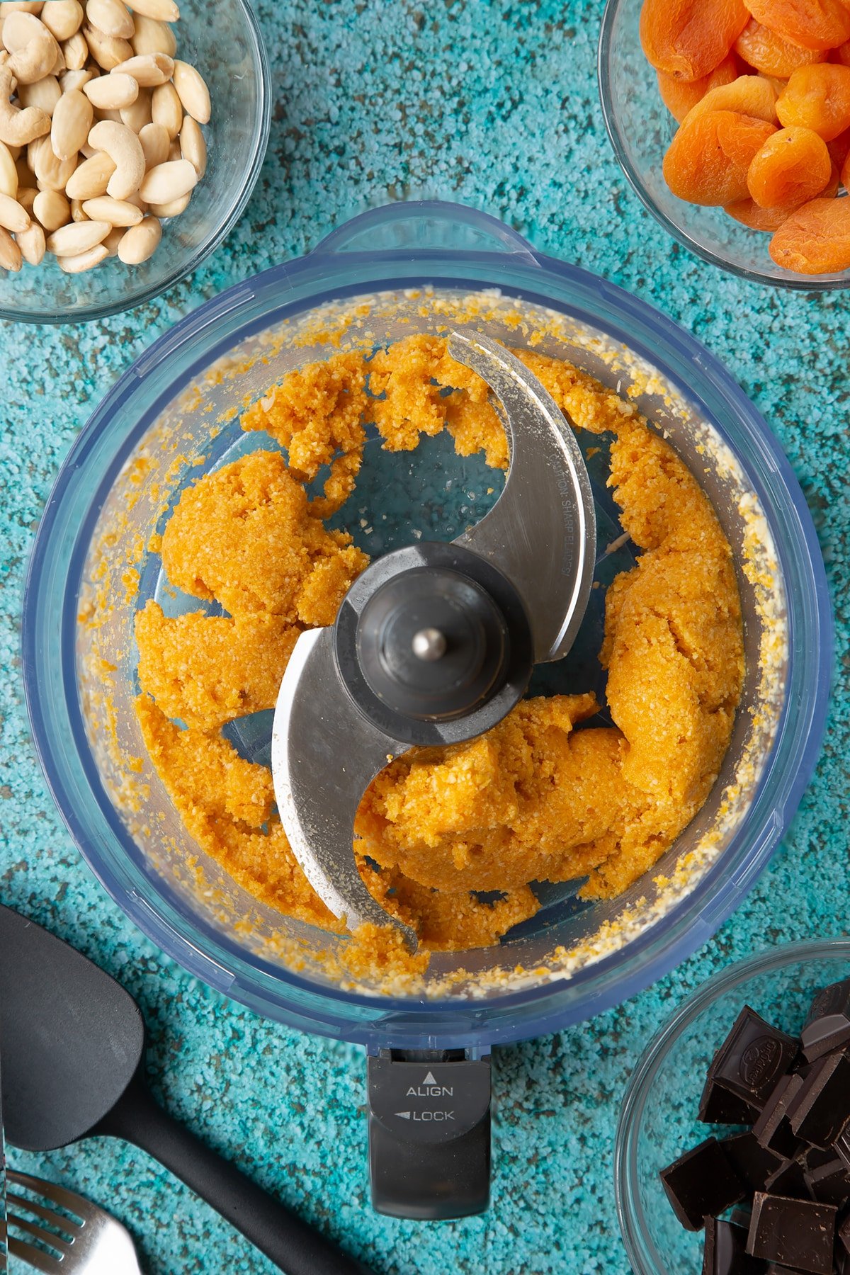 A blender containing almonds, apricots and a few cashews blitzed together to a paste. Ingredients to make chocolate apricot balls surround the blender.