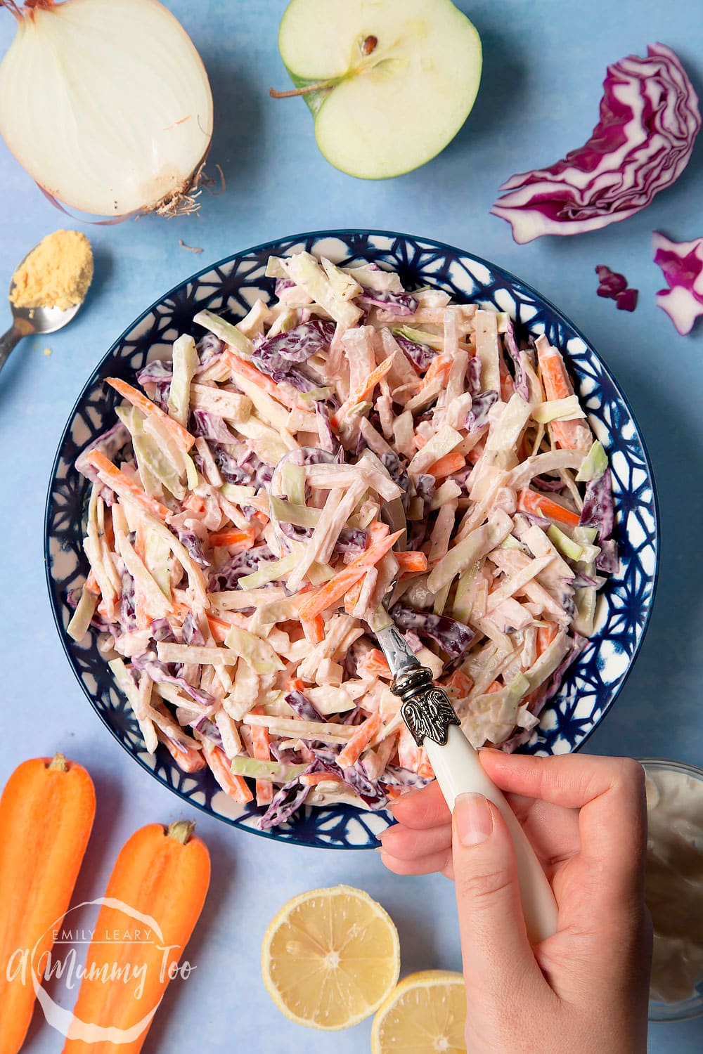coleslaw on a patterened blue bowl with a hand holding a fork surrounded by ingredients on a blue background.