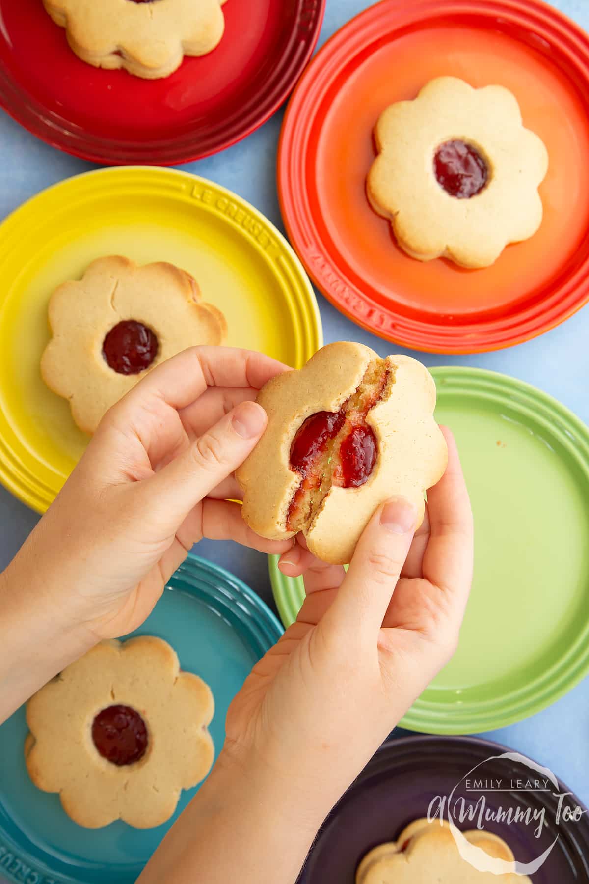 Two hands are holding a giant jammie dodger, broken in half. More jammie dodgers sit on a variety of coloured small plates in the background.