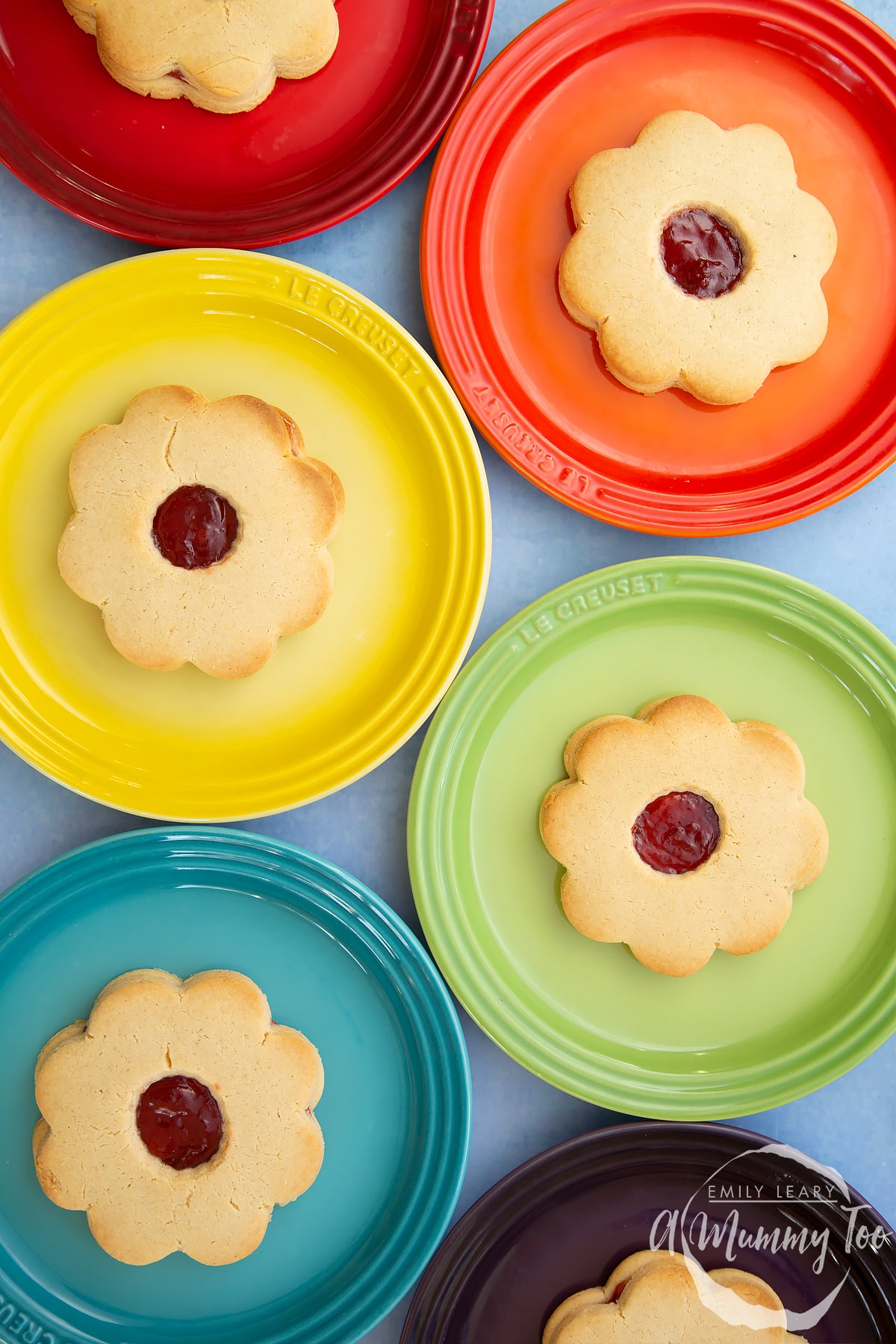 Giant jammie dodgers arranged on a variety of rainbow coloured small plates.