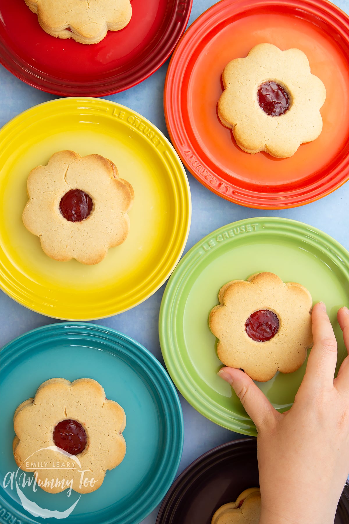 Giant jammie dodgers arranged on a variety of coloured small plates. A hand reaches in to take one from the green plate.