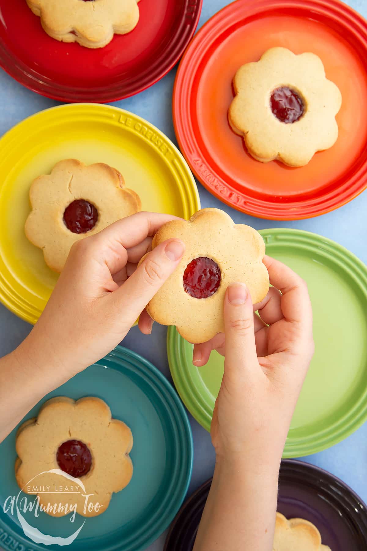 Two hands are holding a giant jammie dodger. More jammie dodgers sit on a variety of coloured small plates in the background.