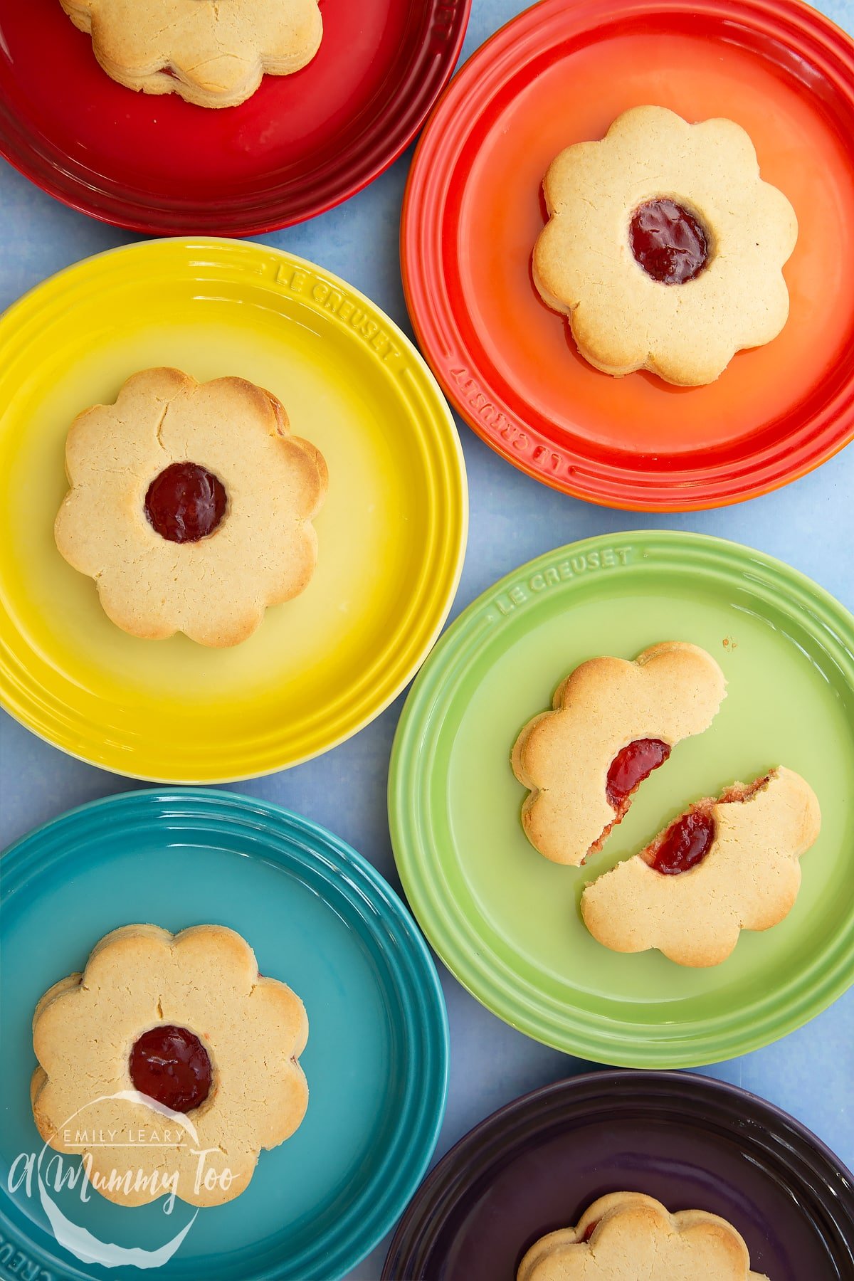 Giant jammie dodgers arranged on a variety of rainbow coloured small plates. One of the jammie dodgers is broken in half.