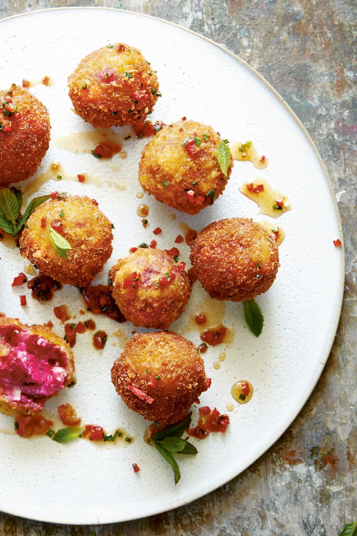 Goat's cheese croquettes on a large white plate. They have been drizzled with maple syrup, red chilli and chive dressing and garnished with mint. One croquette has been opened, showing the pinkish red interior.