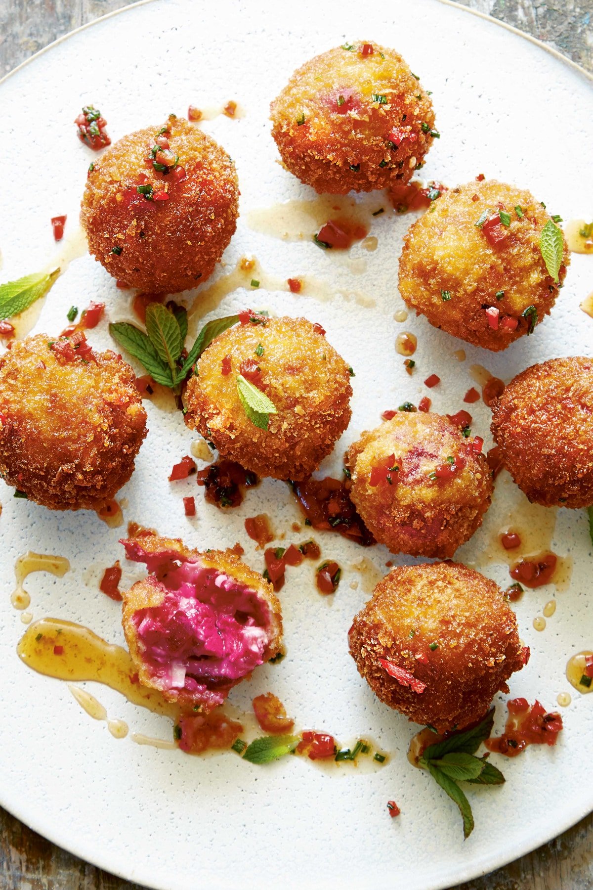 Goat's cheese croquettes on a large white plate. They have been drizzled with maple syrup, red chilli and chive dressing and garnished with mint. 