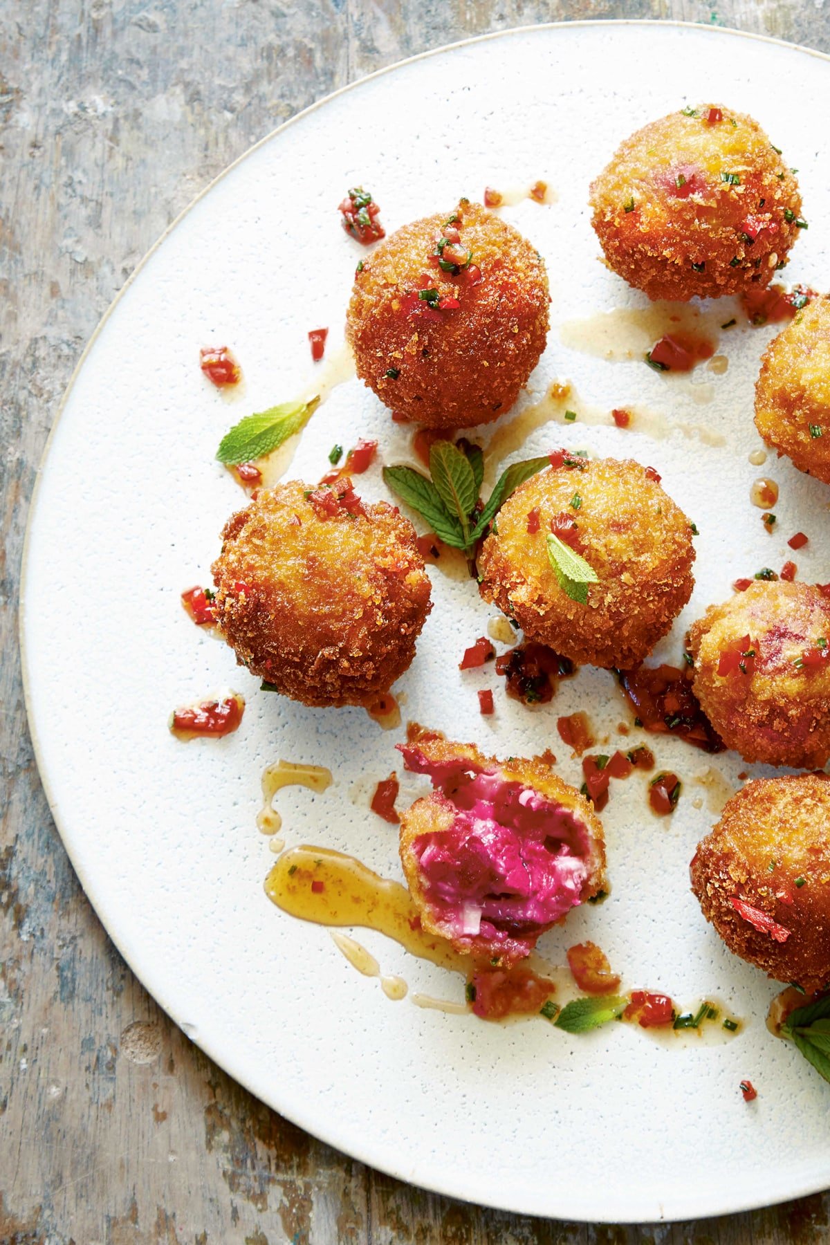 Goat's cheese croquettes on a large white plate. They have been dressed with maple syrup, red chilli, chives and mint. One croquette has been opened, showing the beetroot interior.