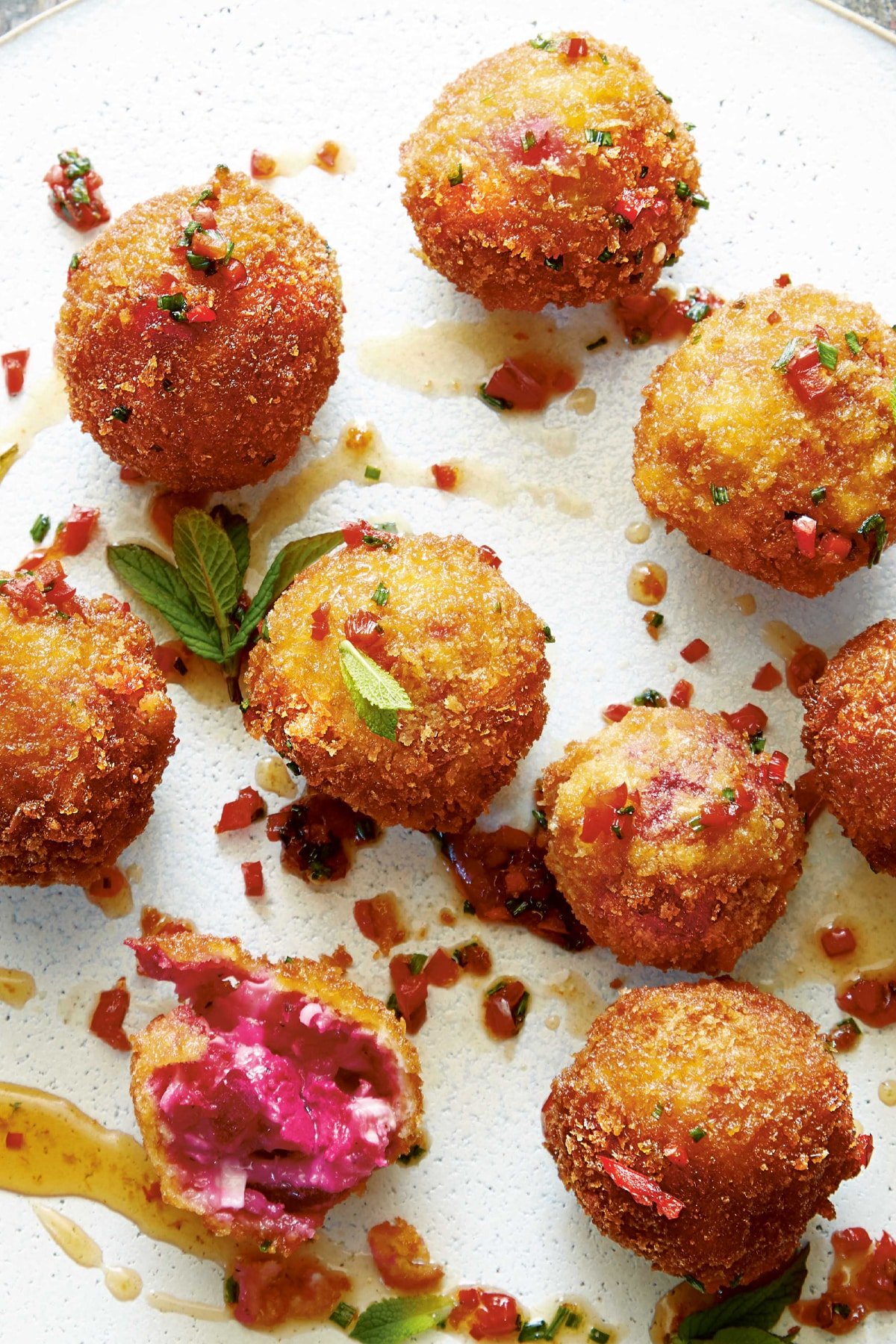 Goat's cheese croquettes on a large white plate. They have been dressed with maple syrup, red chilli, chives and mint. One croquette has been opened, showing the pinkish red interior.