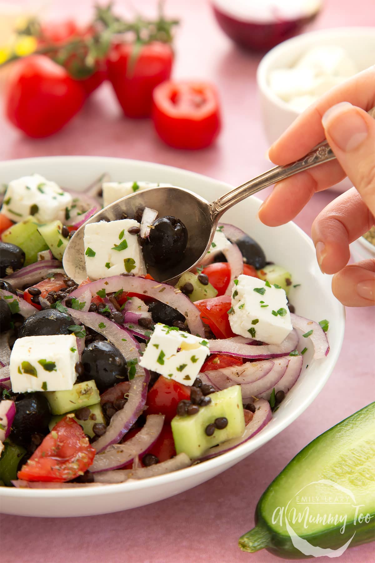 Greek lentil salad in a white serving bowl. The salad is topped with squares of feta and garnished with fresh parsley. A hand delves into the salad with a serving spoon.