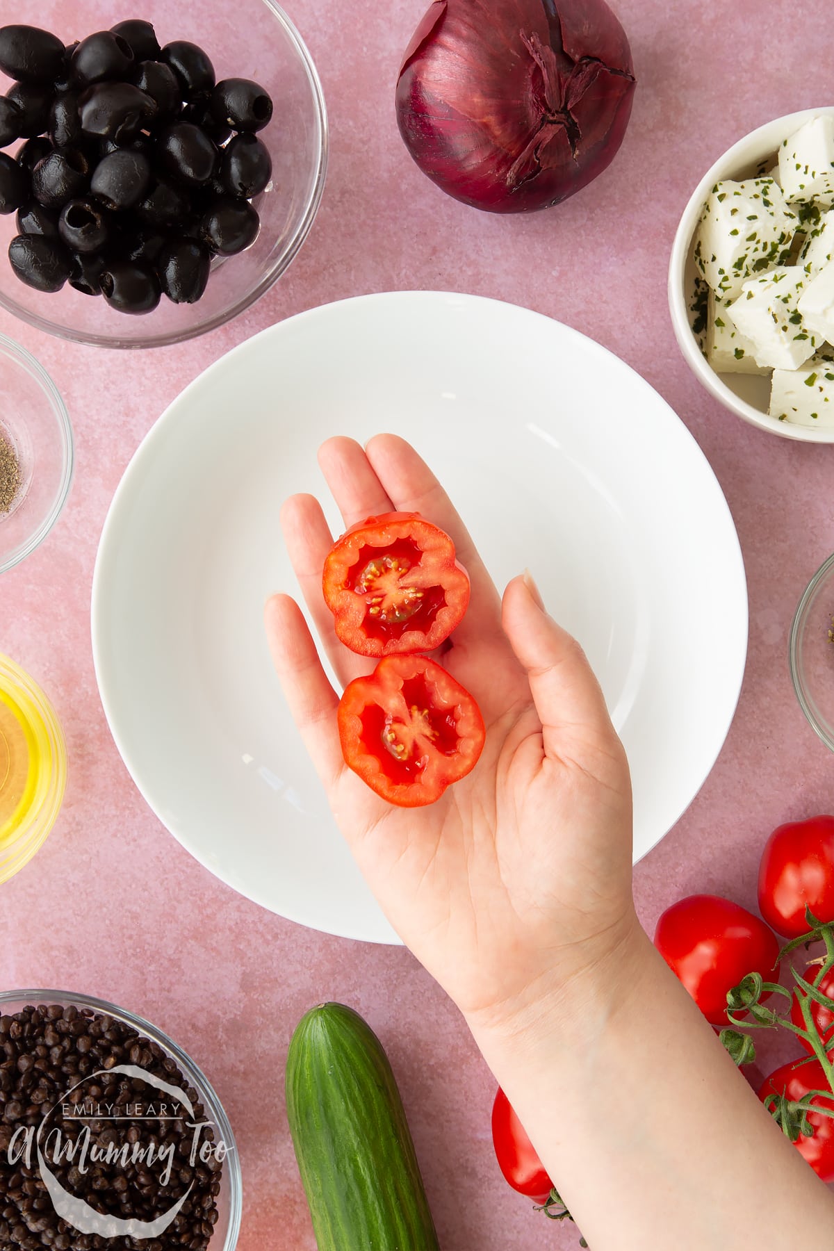 A white bowl with a hand over it holding a tomato cut in half. Ingredients to make Greek lentil salad surround the bowl.