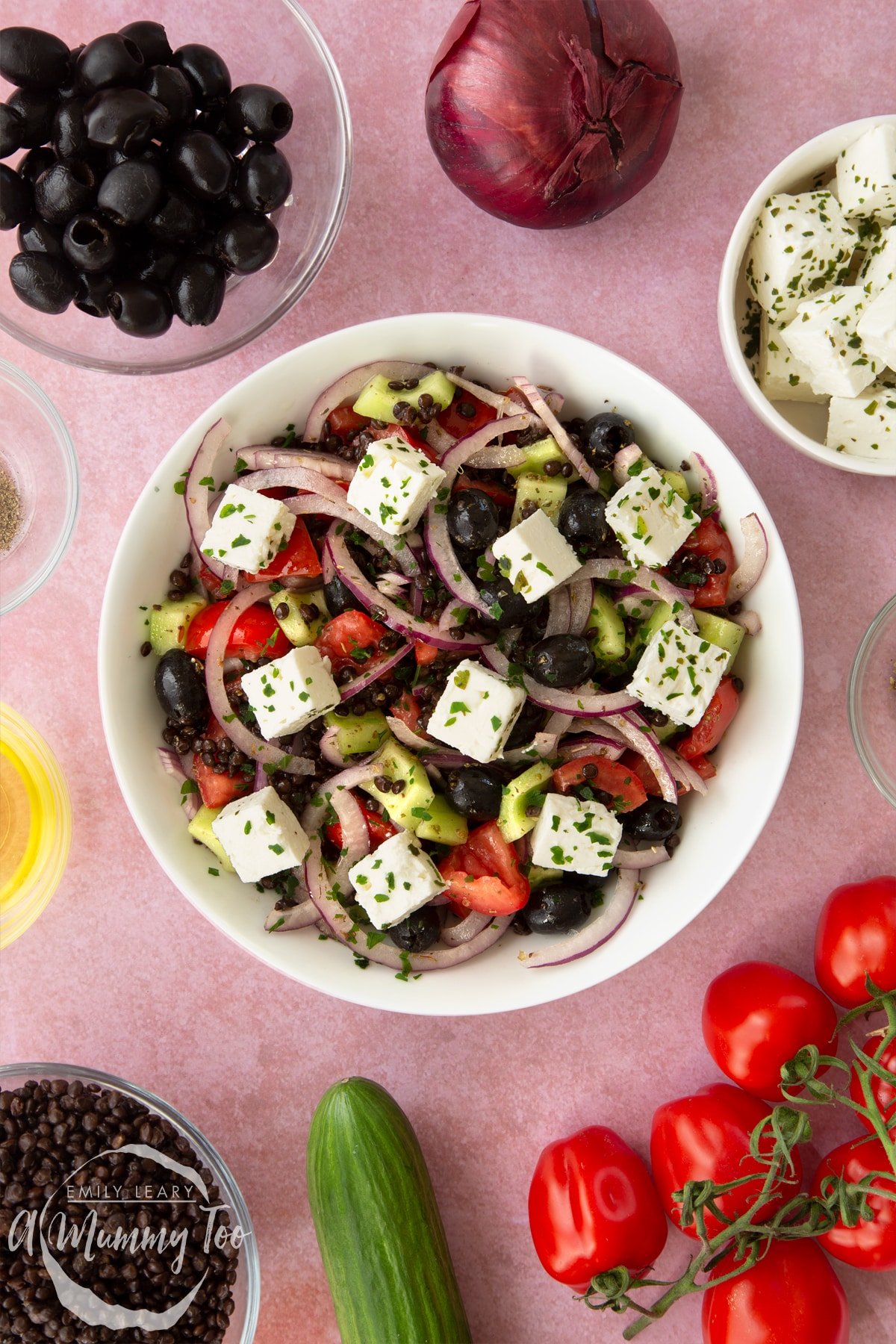 Greek lentil salad in a white serving bowl. Ingredients to make the salad surround the bowl, such as tomatoes, olives, lentils and cucumber.