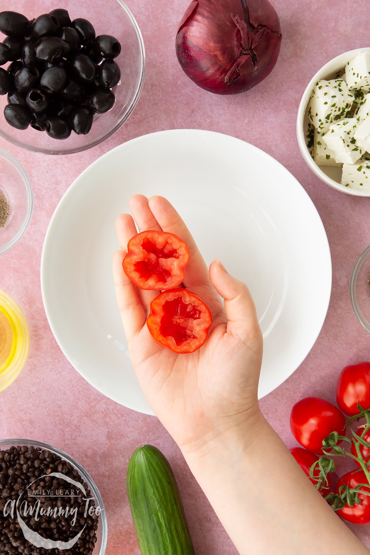 A white bowl with a hand over it holding a tomato cut in half with the seeds removed. Ingredients to make Greek lentil salad surround the bowl.