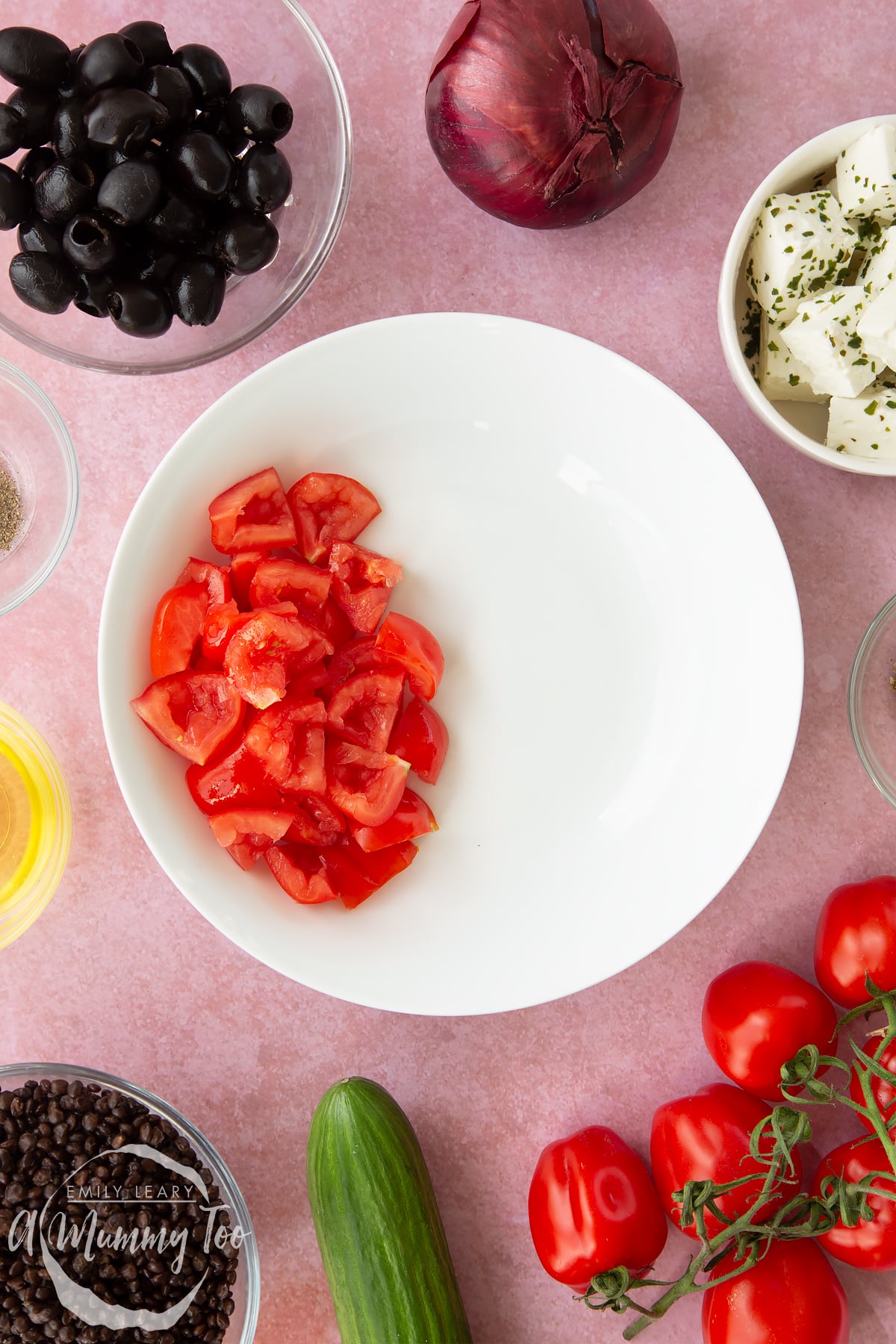 A white bowl containing chopped tomatoes. Ingredients to make Greek lentil salad surround the bowl.