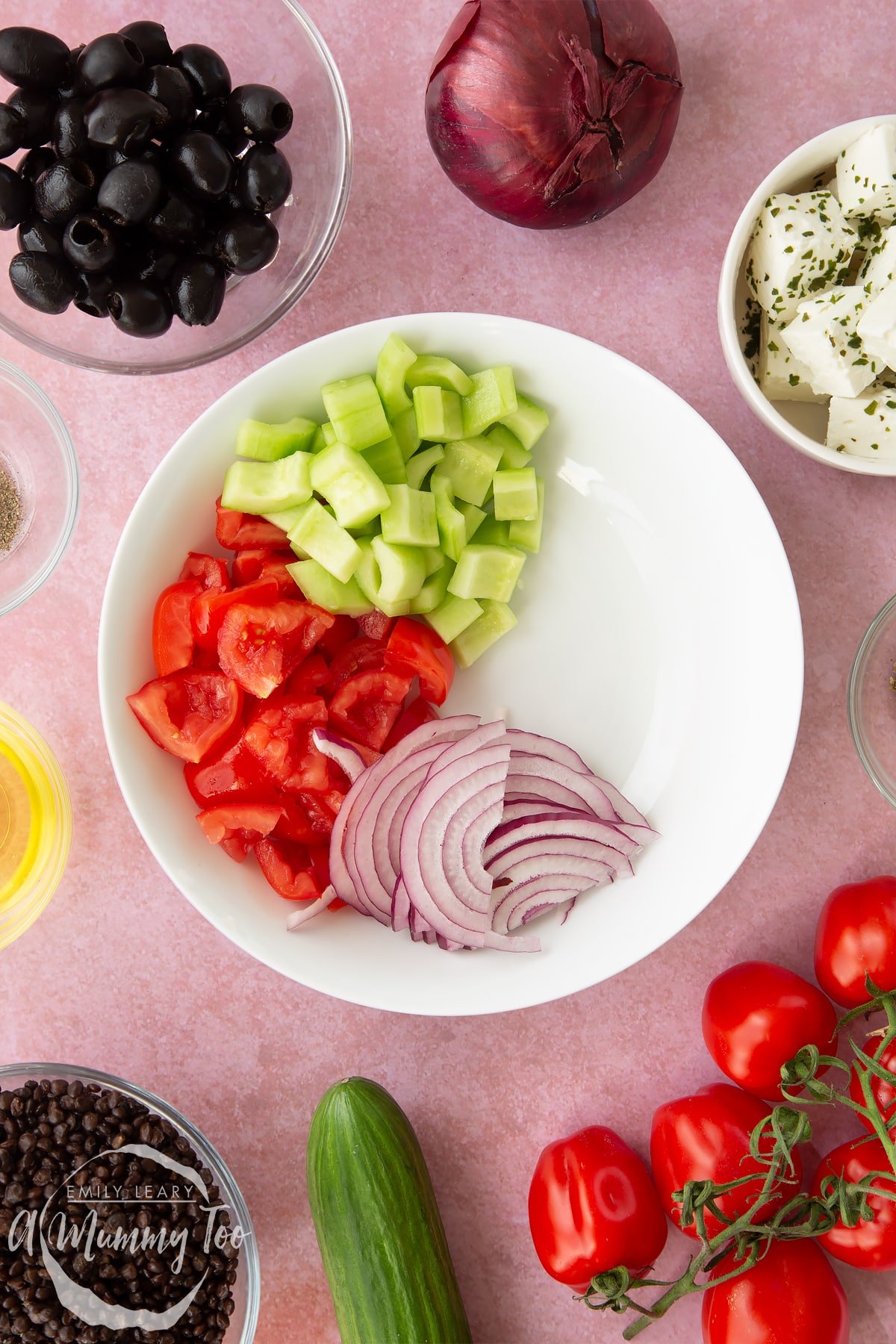 A white bowl containing chopped tomatoes, chopped cucumber and sliced red onion. Ingredients to make Greek lentil salad surround the bowl.