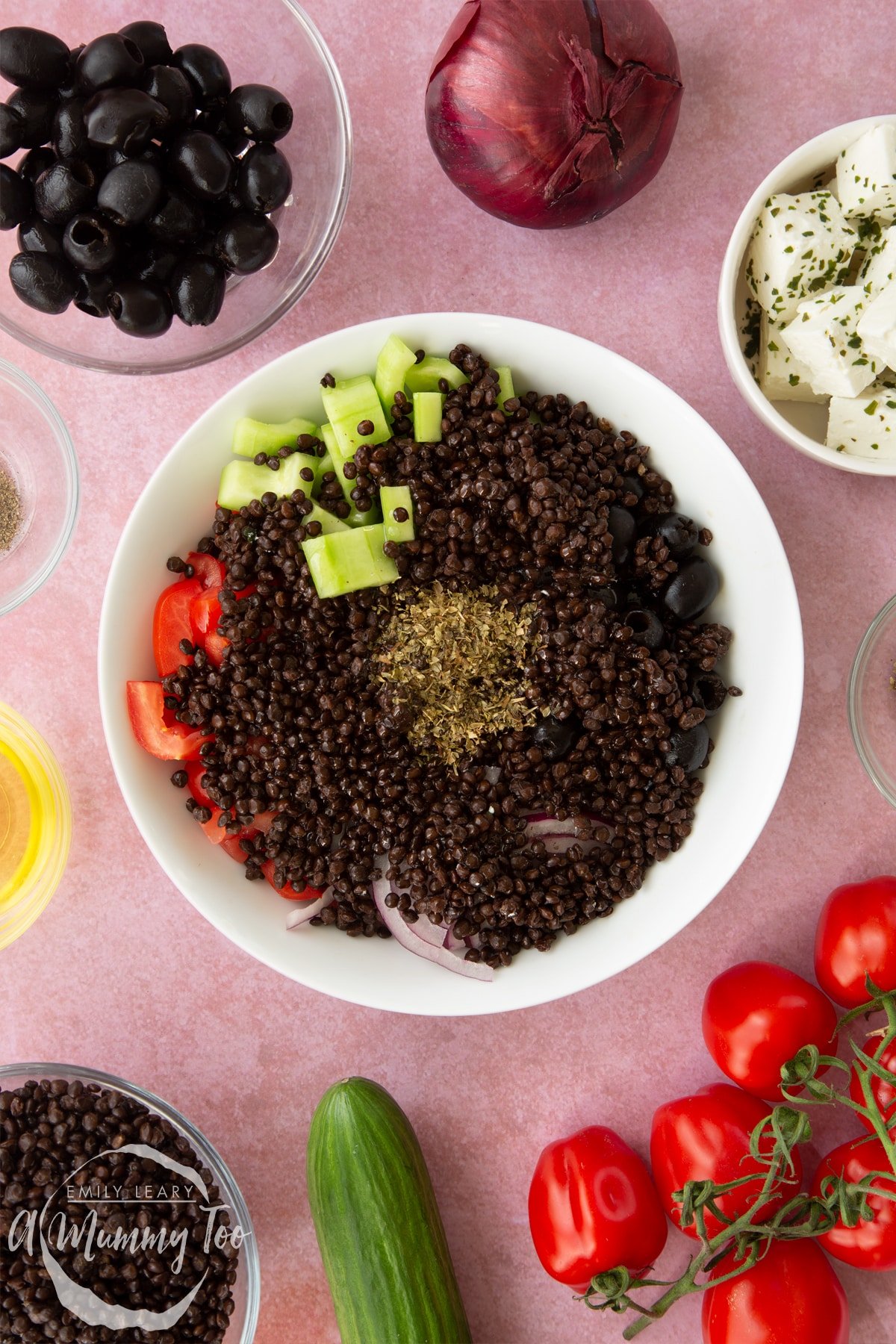 A white bowl containing chopped tomatoes, chopped cucumber, sliced red onion, whole black olives, cooked lentils, olive oil and oregano. Ingredients to make Greek lentil salad surround the bowl.