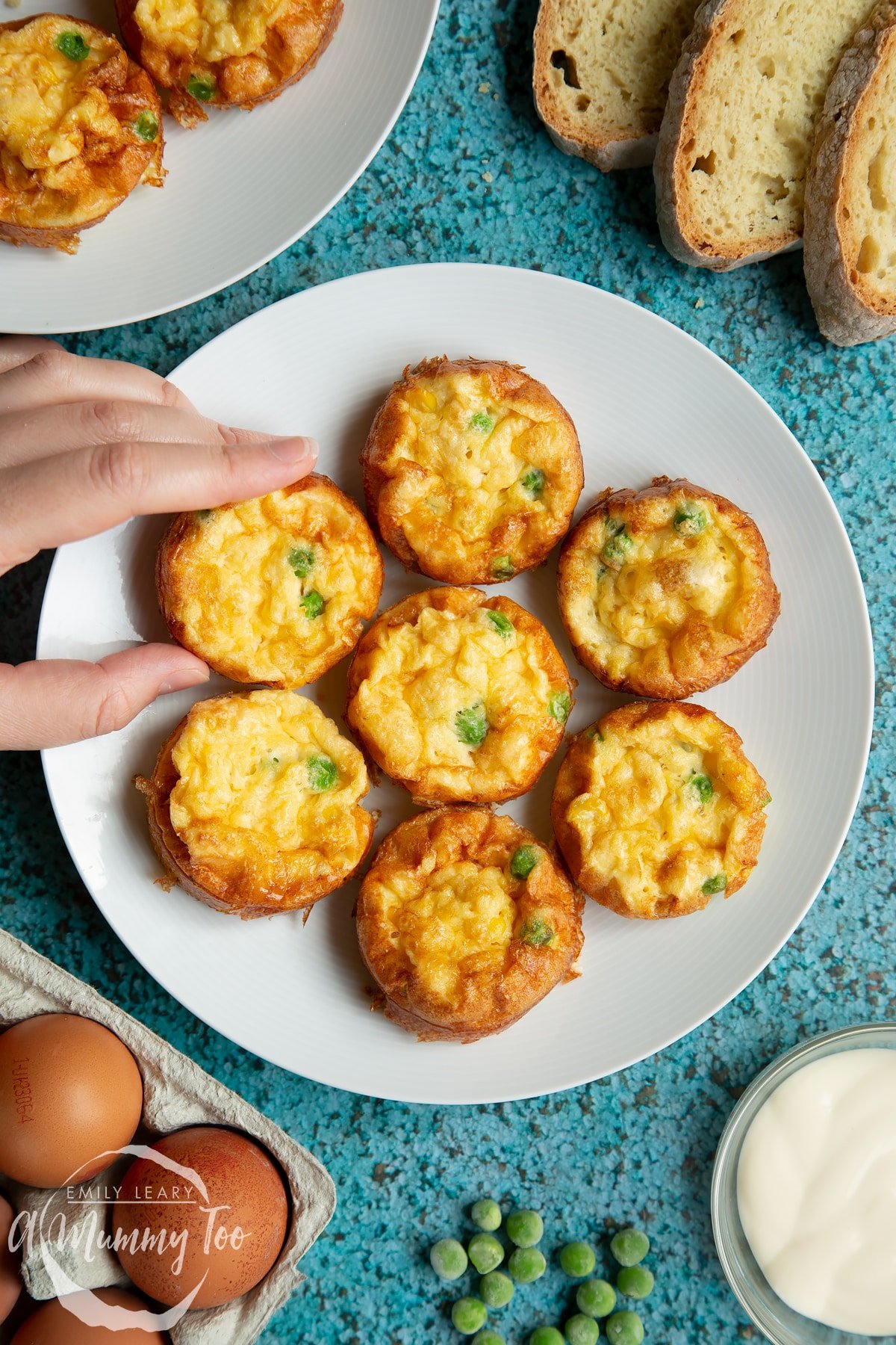 Mini vegetable frittatas arranged on a white plate. Slices of bread and ingredients surround the plate. A hand reaches in to take a frittata.