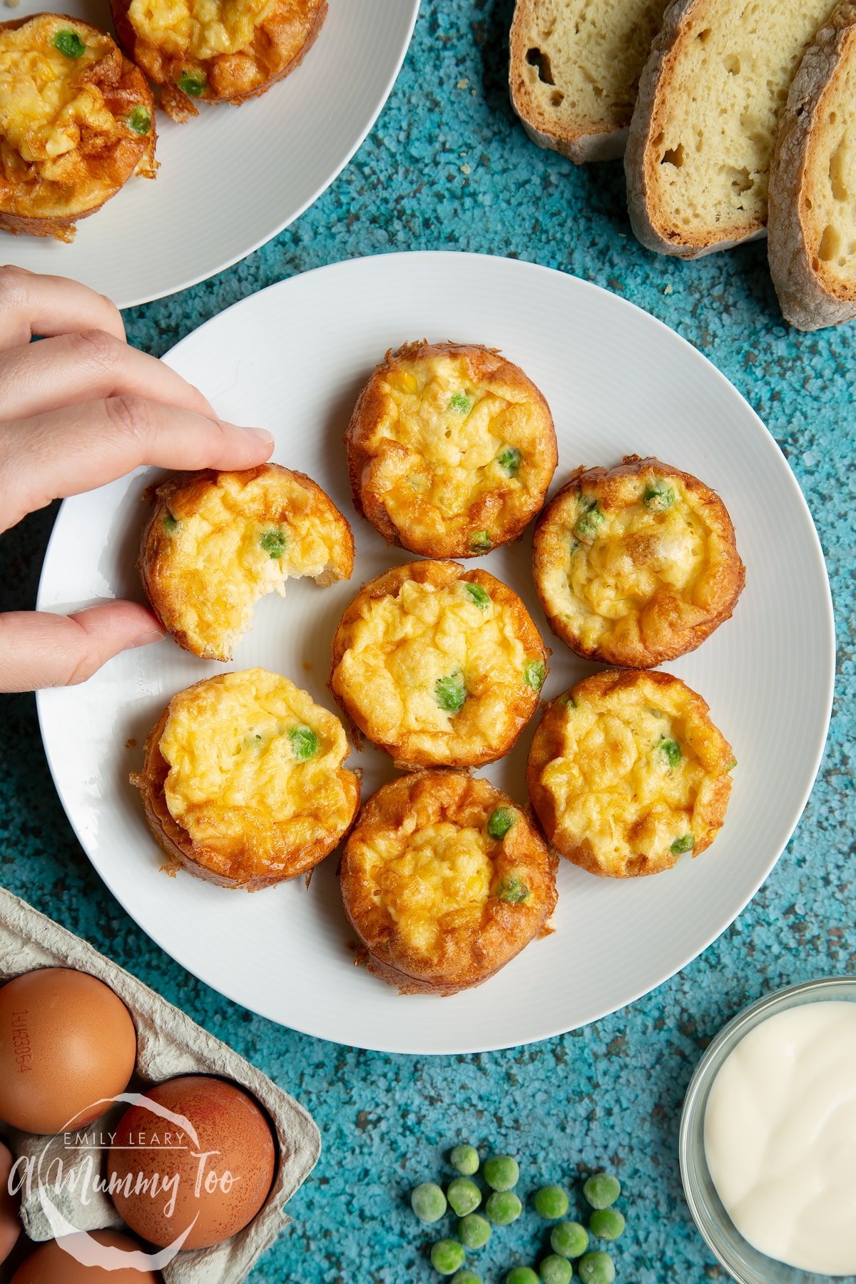 Mini vegetable frittatas arranged on a white plate. Slices of bread and ingredients surround the plate. A hand reaches in to take a frittata, which has a bite taken out of it.