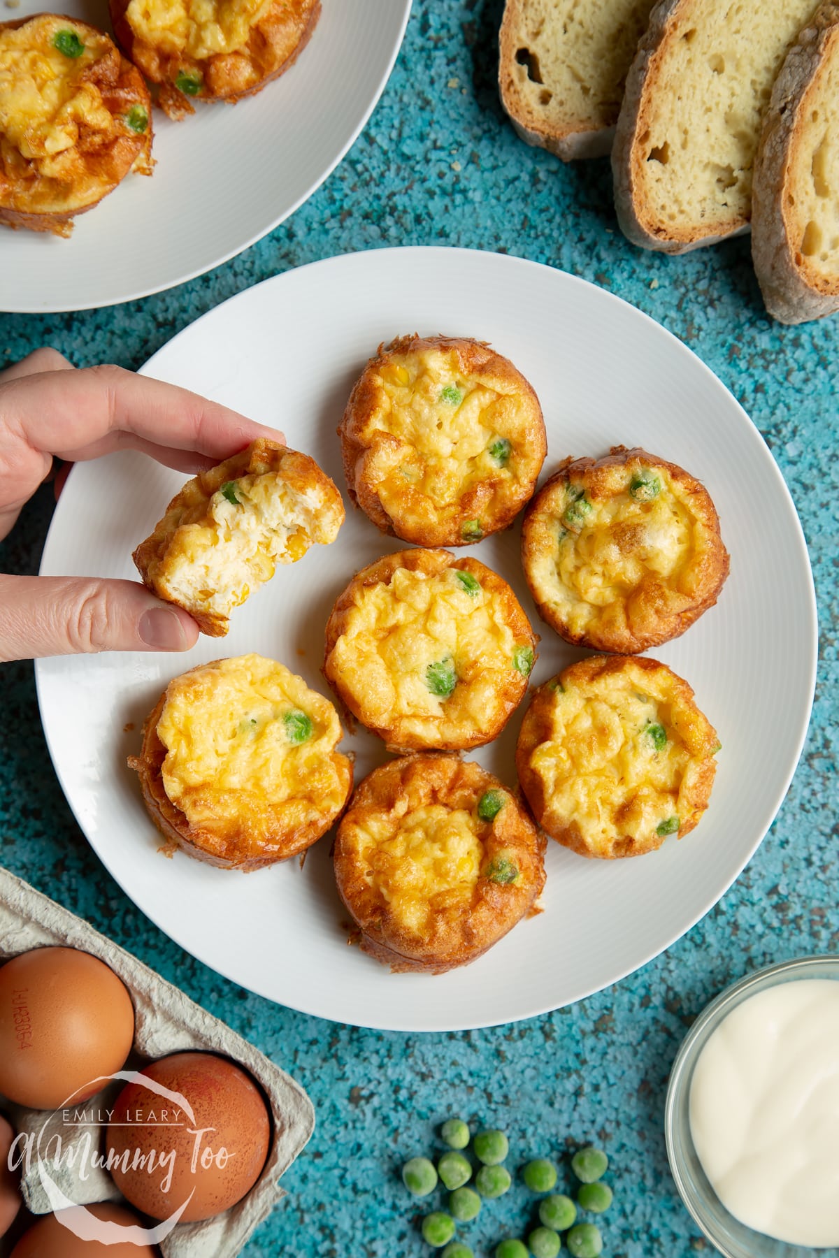 Mini vegetable frittatas arranged on a white plate. Slices of bread and ingredients surround the plate. A hand reaches in to take a frittata, tilted to show the creamy, fluffy inner.