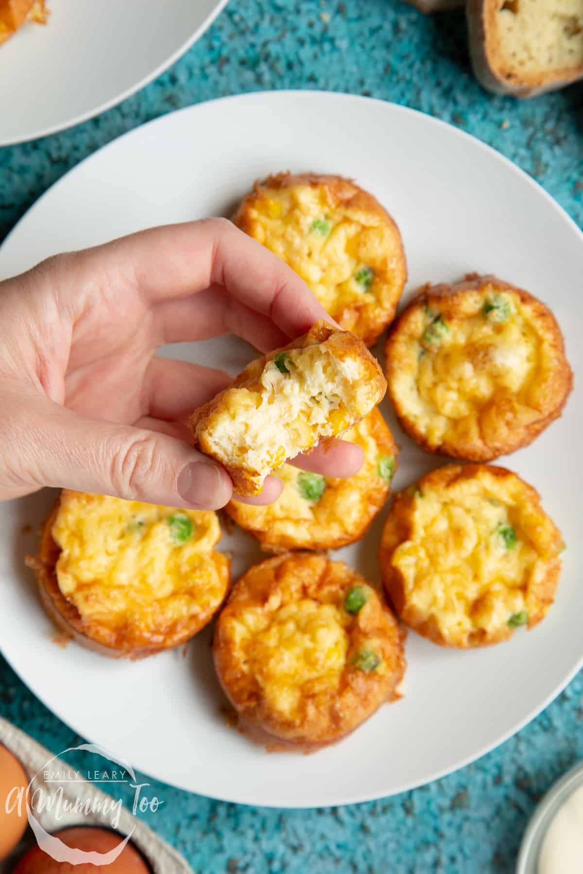 A holding a frittata, tilted to show the creamy, fluffy inner. In the background, more mini vegetable frittatas are arranged on a white plate. 