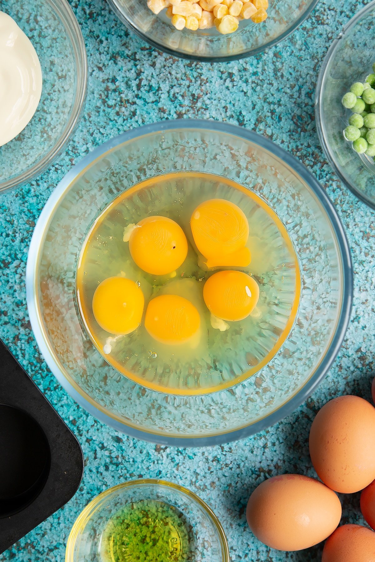Eggs in a mixing bowl. Ingredients to make mini vegetable frittatas surround the bowl.