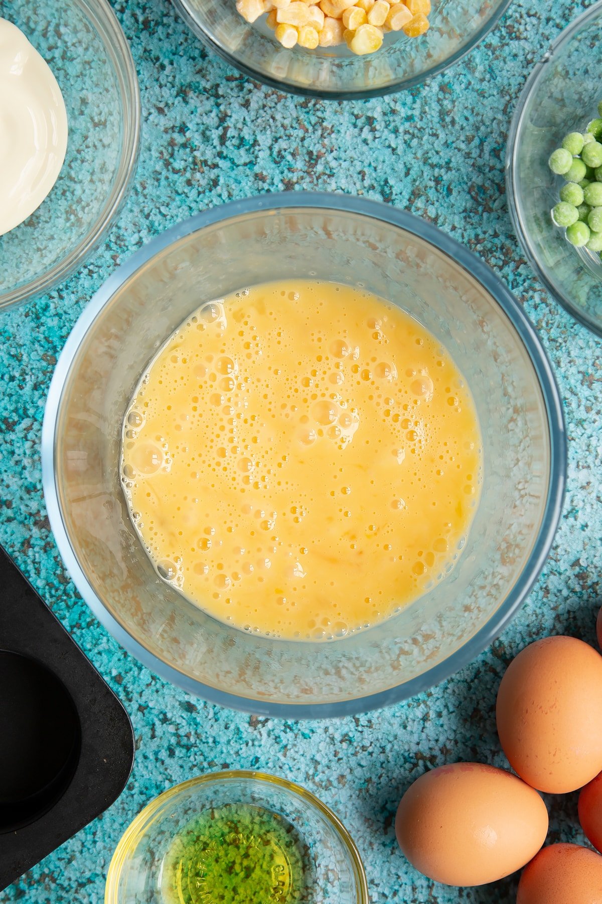 Beaten eggs in a mixing bowl. Ingredients to make mini vegetable frittatas surround the bowl.