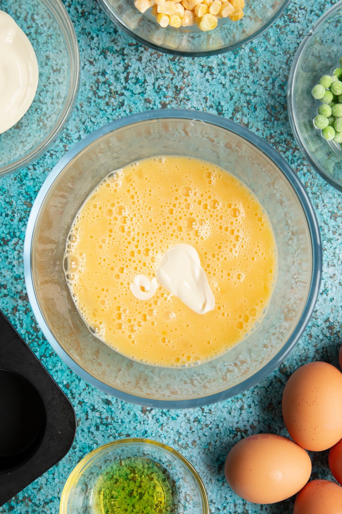 Beaten eggs in a mixing bowl with cream cheese added. Ingredients to make mini vegetable frittatas surround the bowl.