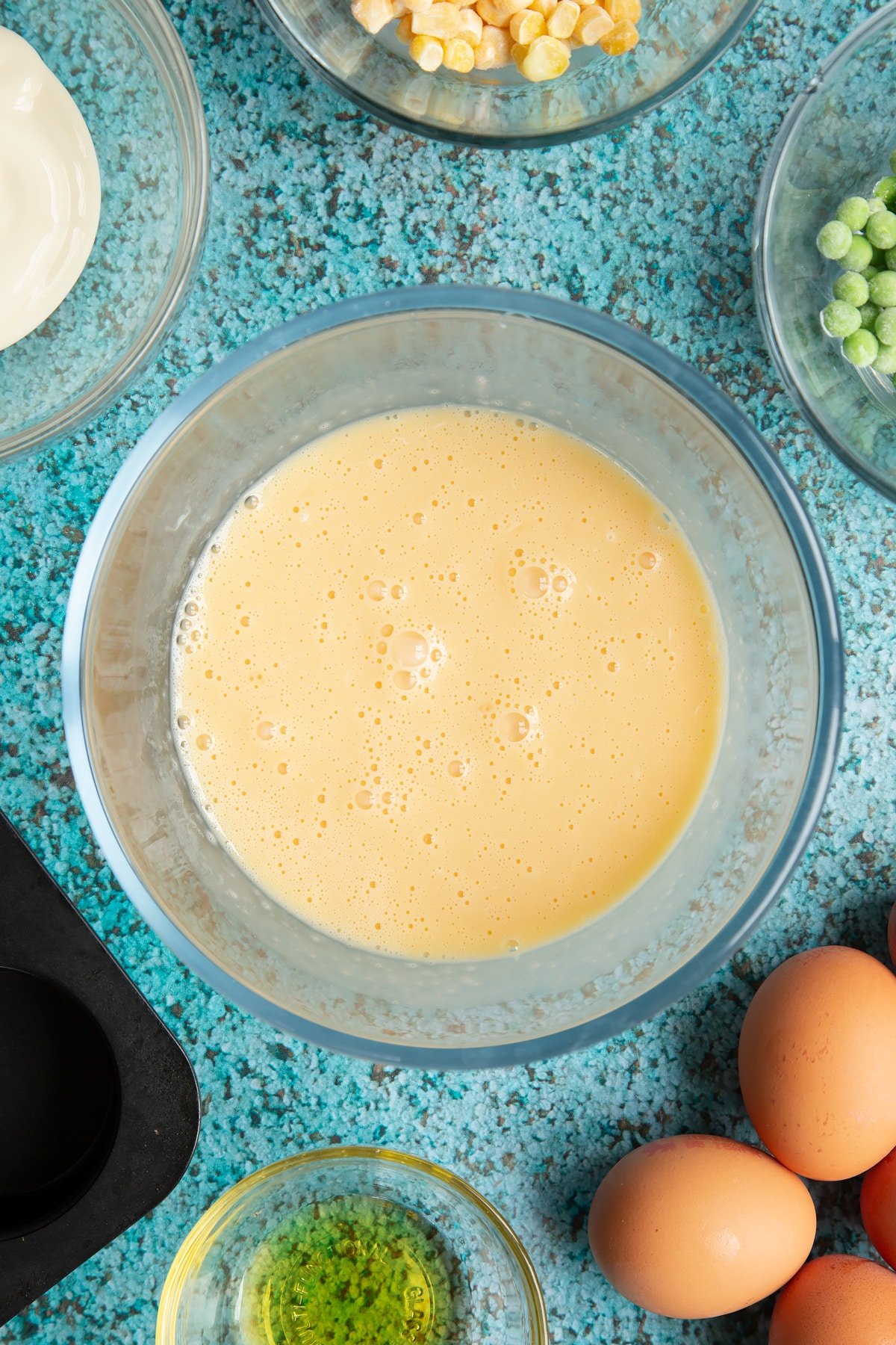 Eggs and cream cheese beaten together in a mixing bowl. Ingredients to make mini vegetable frittatas surround the bowl.