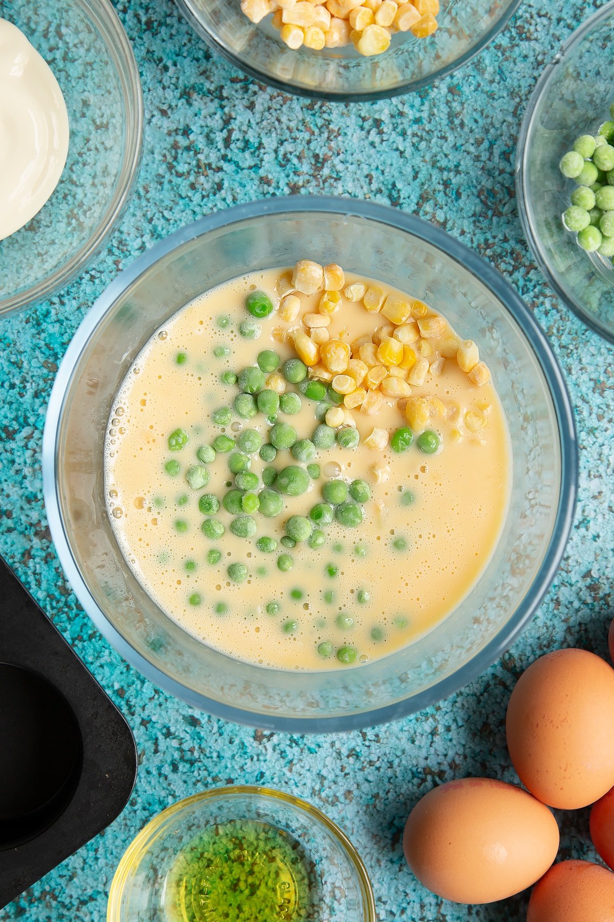 Eggs and cream cheese beaten together in a mixing bowl. Peas and sweetcorn are shown on top. Ingredients to make mini vegetable frittatas surround the bowl.