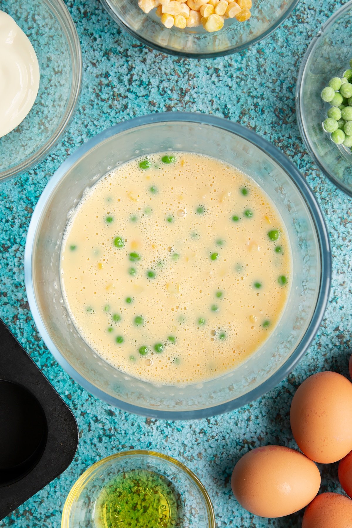 Eggs and cream cheese beaten together with peas and sweetcorn in a mixing bowl. Ingredients to make mini vegetable frittatas surround the bowl.