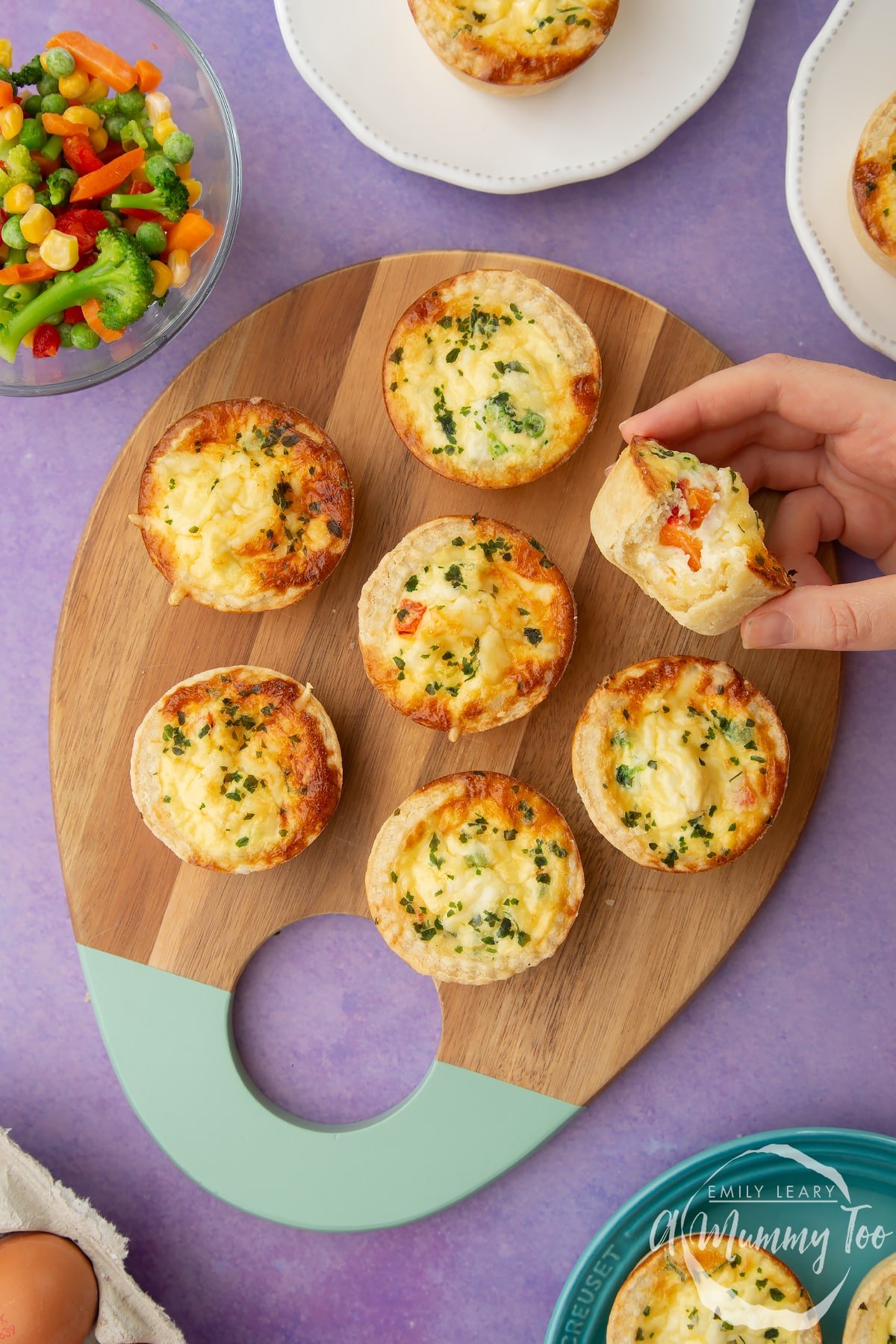 Mini vegetable quiches arranged on a wooden board. A hand holds ones with a bite taken out of it.