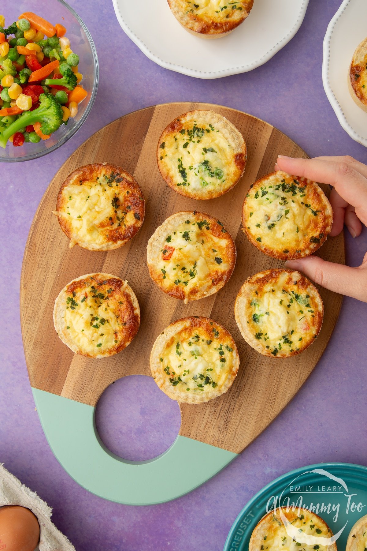 Mini vegetable quiches arranged on a wooden board. A hand reaches to take one.