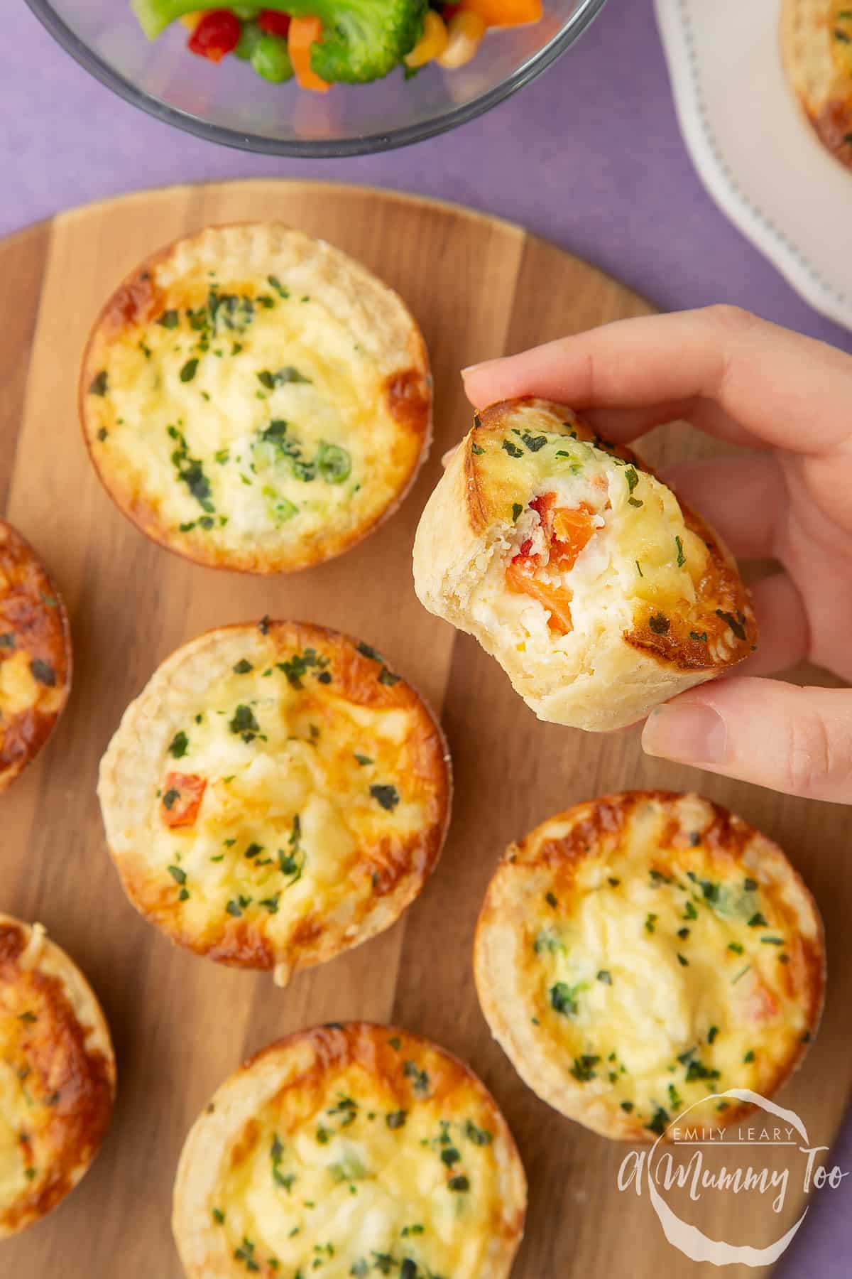 A hand holds a mini vegetable quiche with a bite taken out of it. More mini vegetable quiches are arranged on a wooden board in the background.