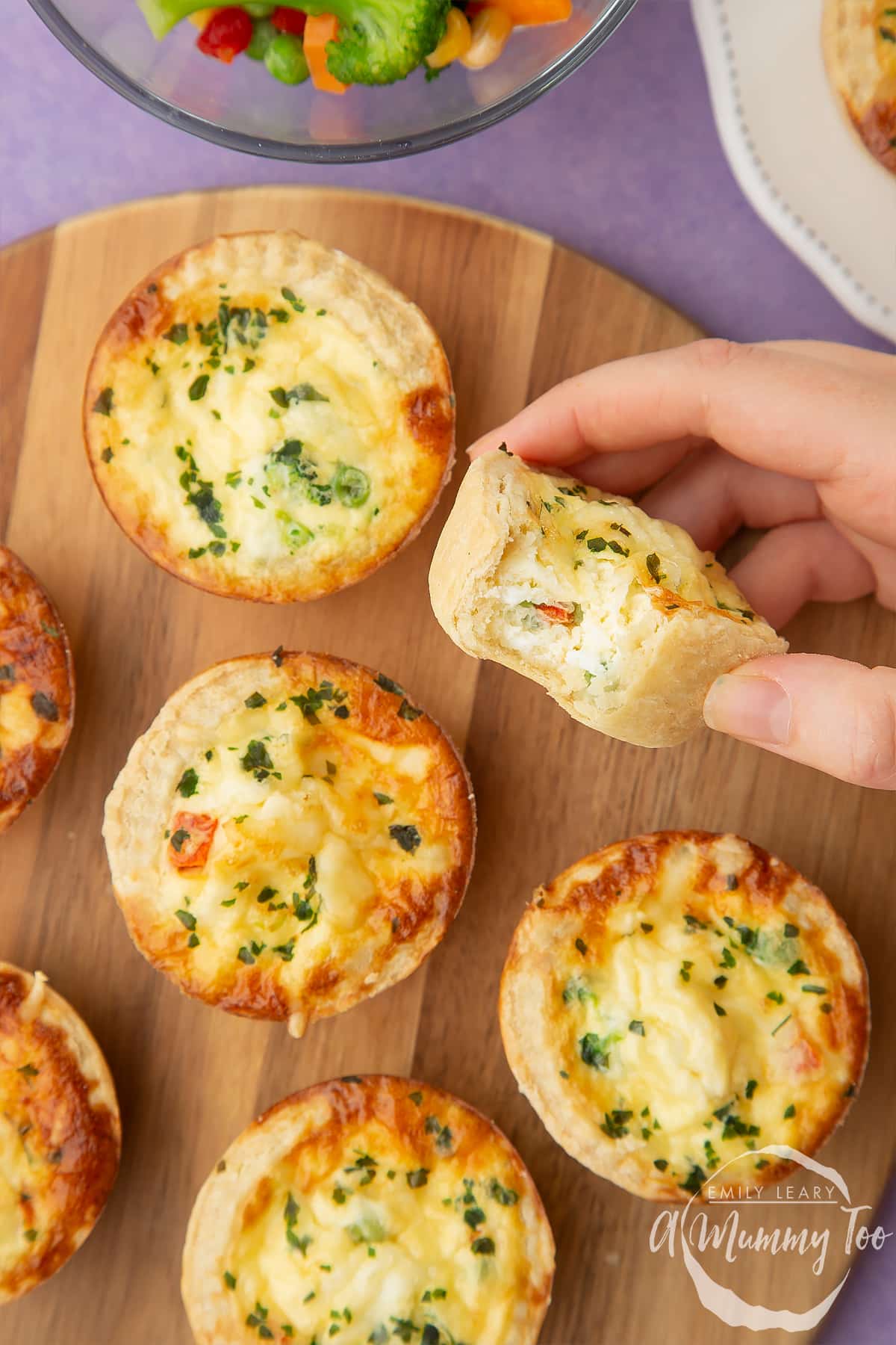 A hand holds a mini vegetable quiche with a bit taken out of it, above more arranged on a wooden board.